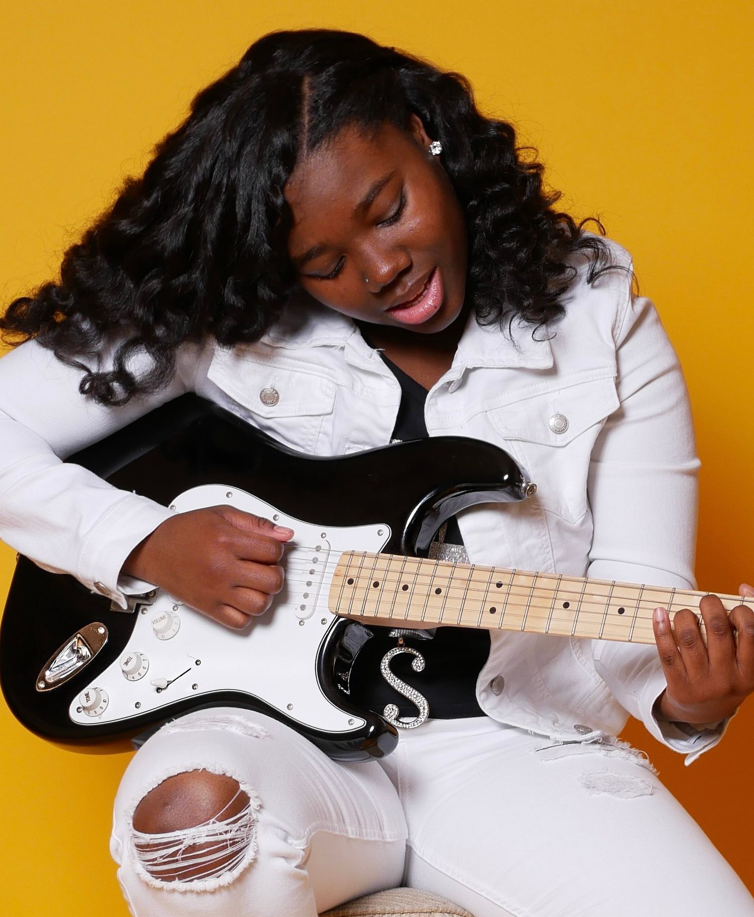 Woman in white denim ensemble plays electric guitar against yellow backdrop.