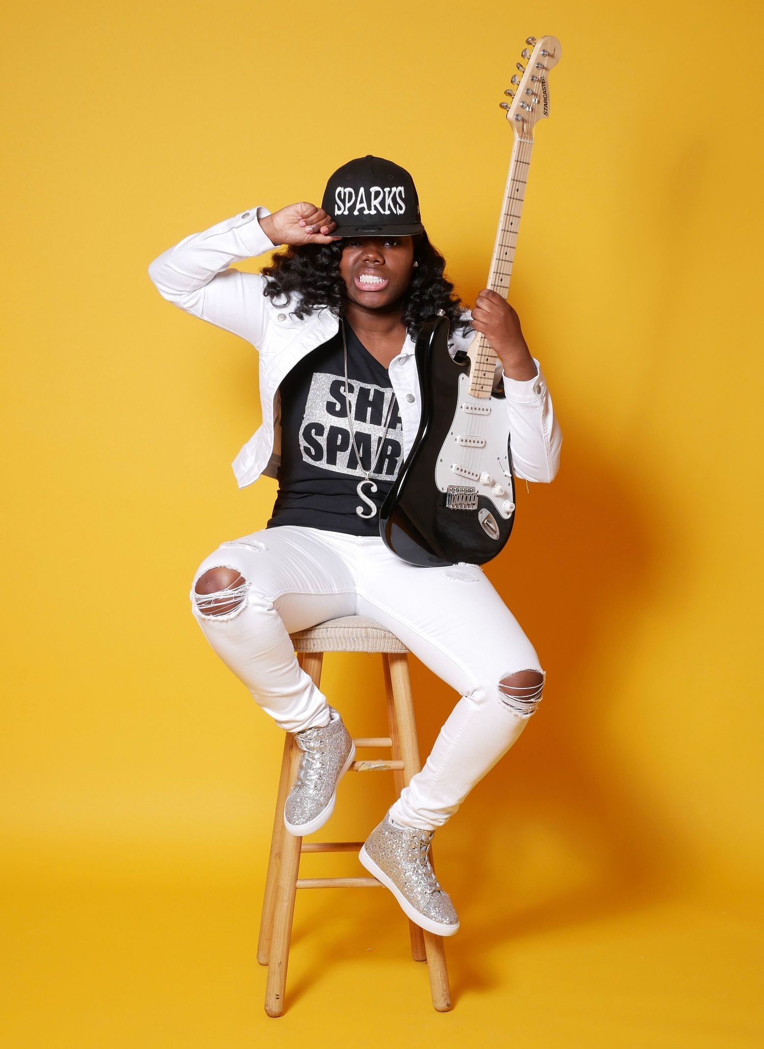 Musician in white ripped jeans and cap poses with electric guitar on stool.