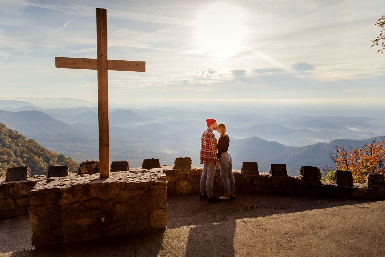 A couple shares an intimate moment near a large cross overlooking a mountainous landscape at sunset.