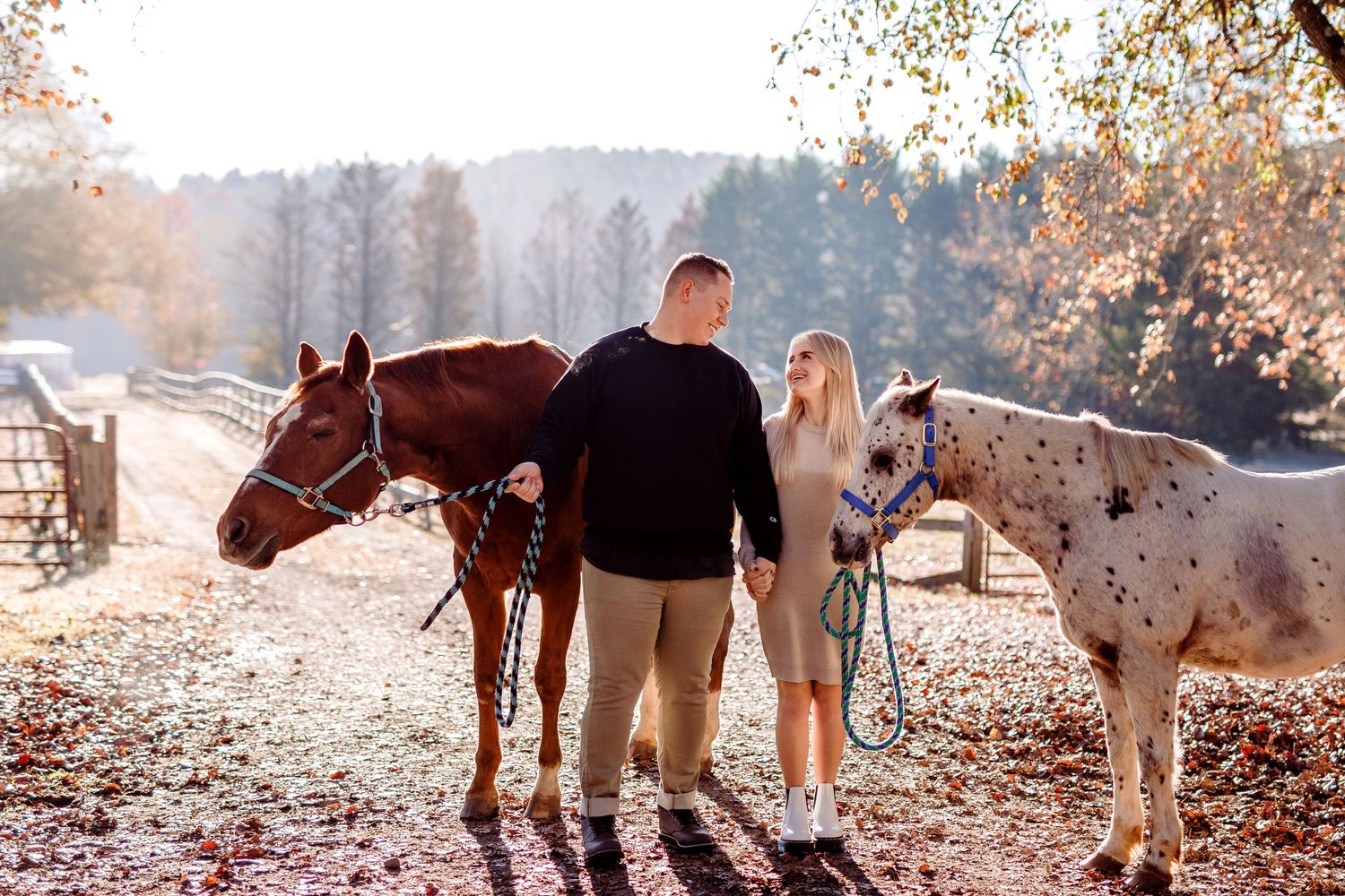 A couple stands with their two horses in a sunlit autumn field.