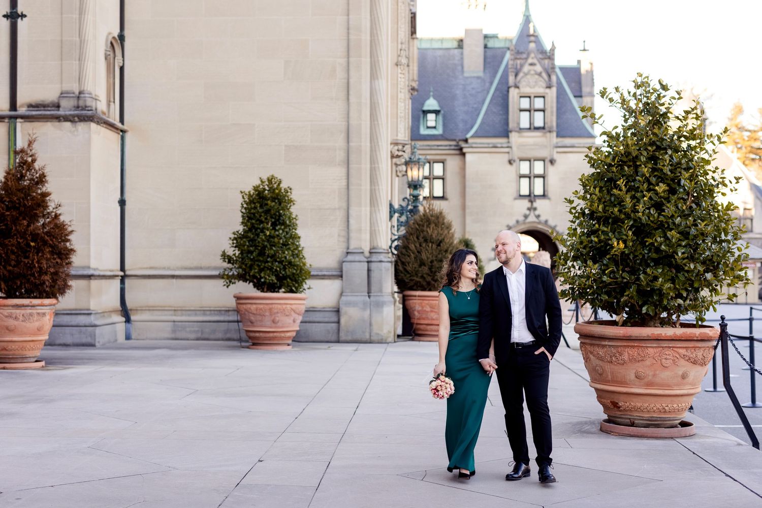A couple strolls along an elegant terrace with potted trees and classical architecture in the background.