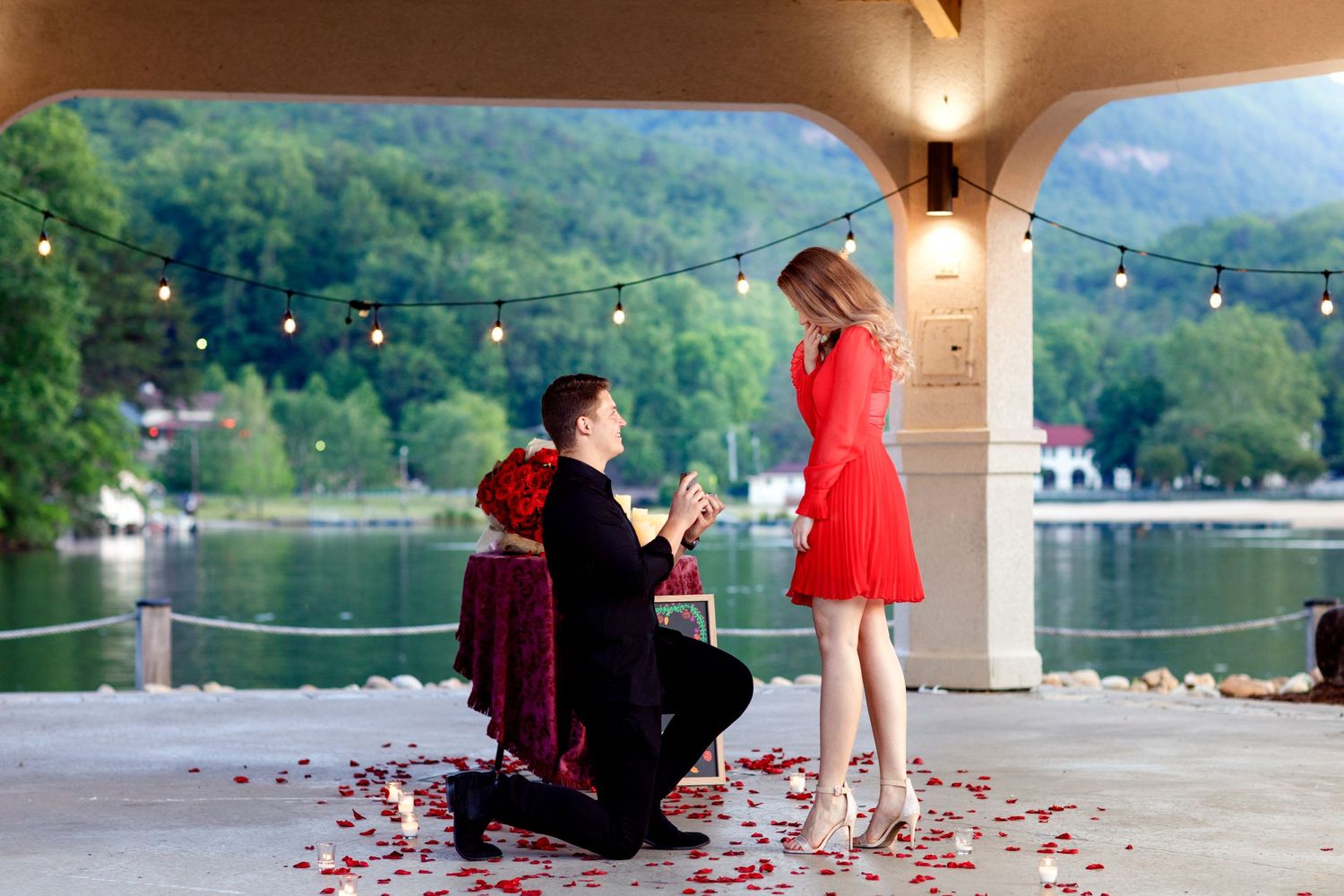 A romantic proposal scene with rose petals scattered under an archway by a lakeside.