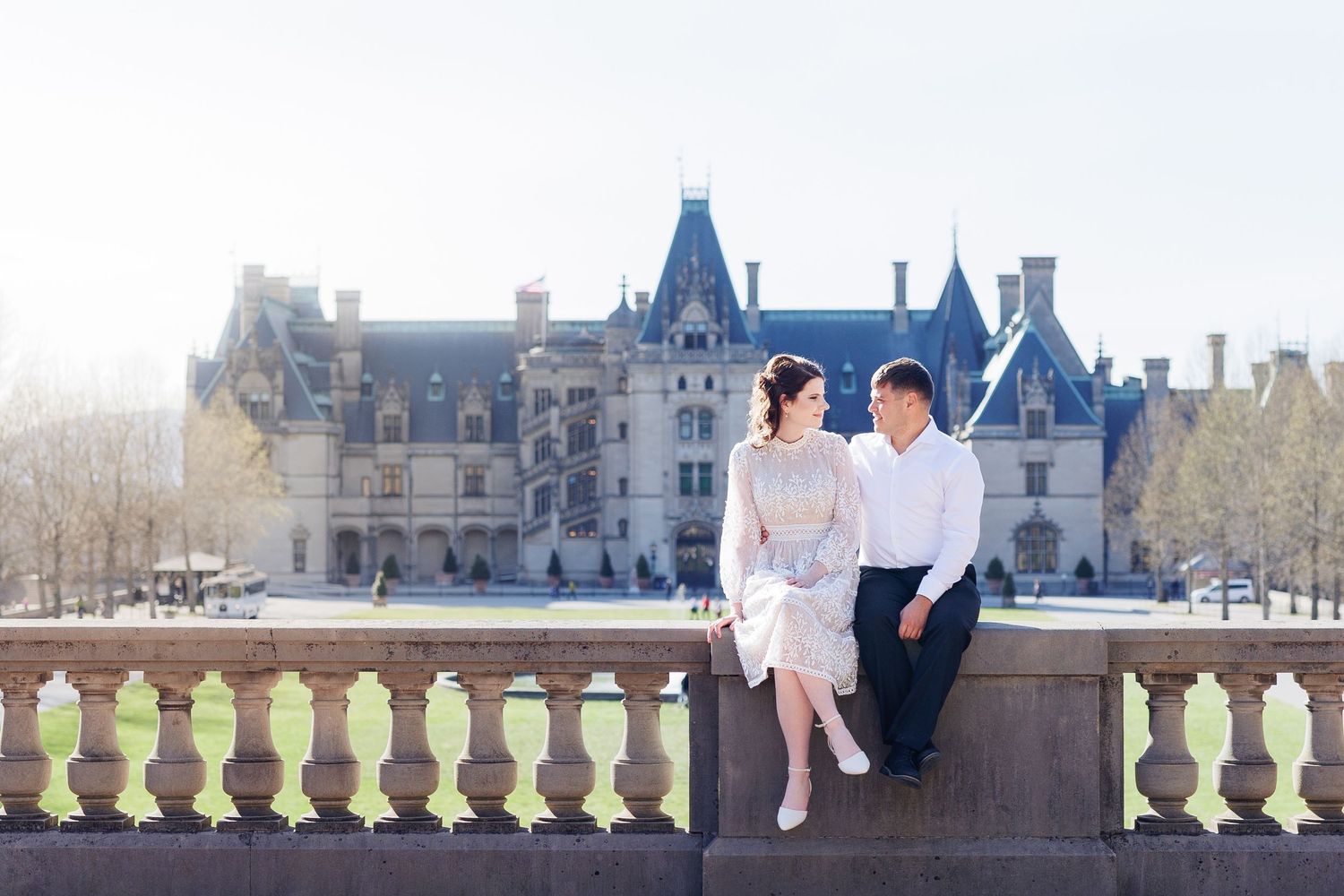 A couple poses romantically on a balcony with a grand castle in the background.