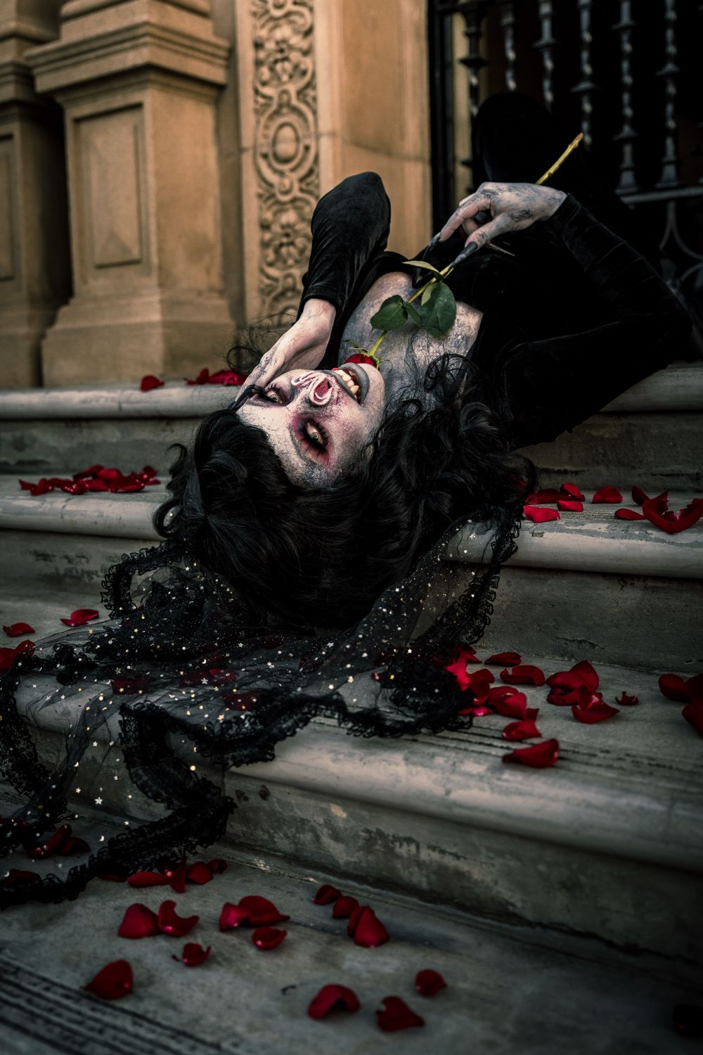 Female model in a haunting yet beautiful bat-inspired horror shoot, lying upside down on steps surrounded by rose petals.