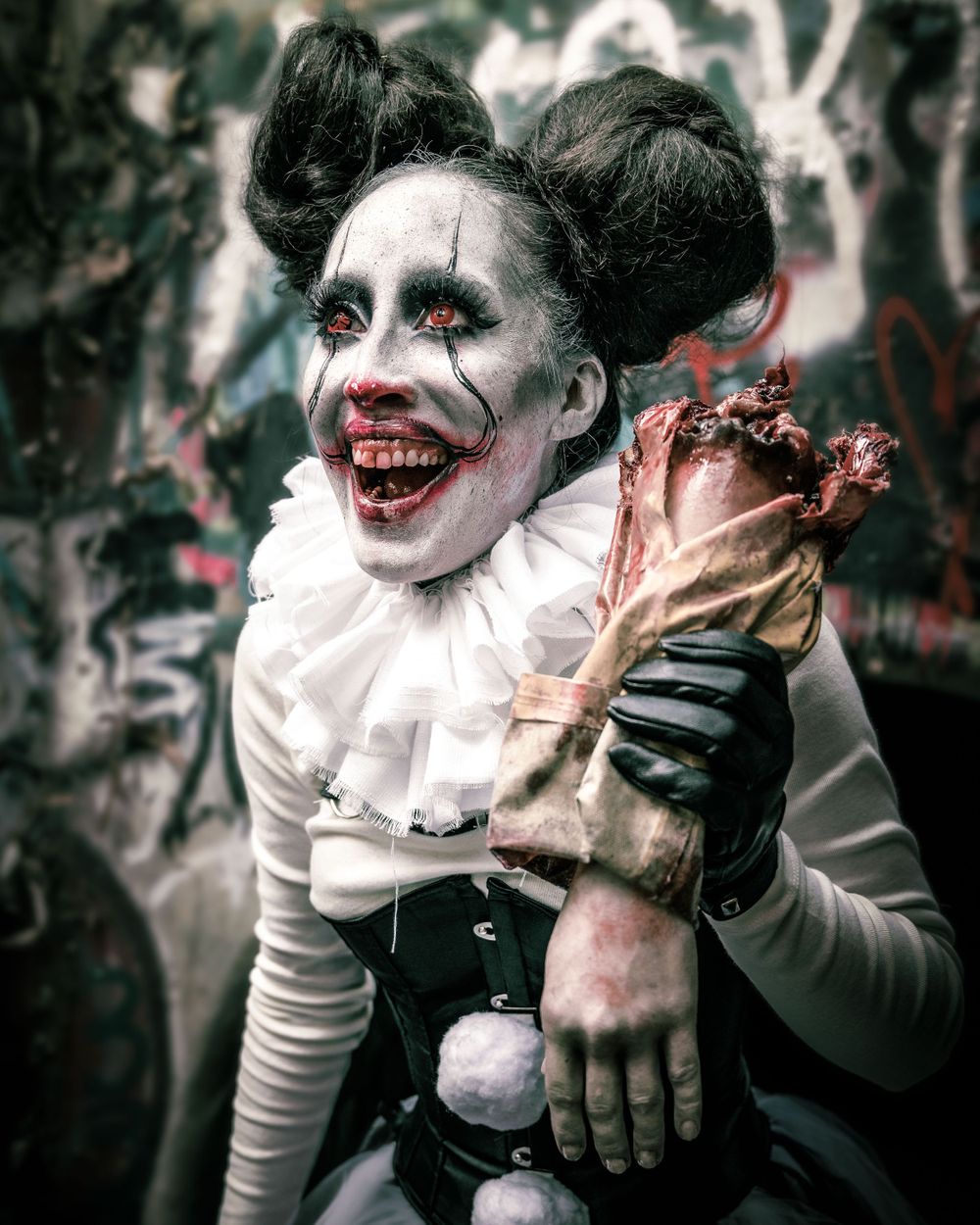 Female model as Pennywise the clown in a graffiti-covered sewer, holding a severed hand prop with red eyes and subtle blood effects.