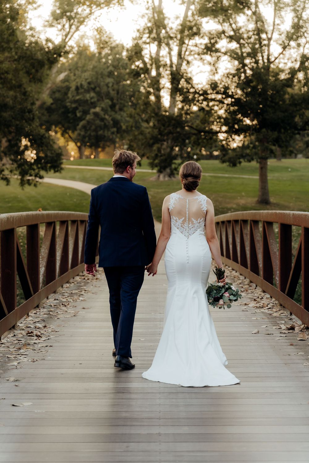 Bride and groom walking away, hand-in-hand, across a bridge.