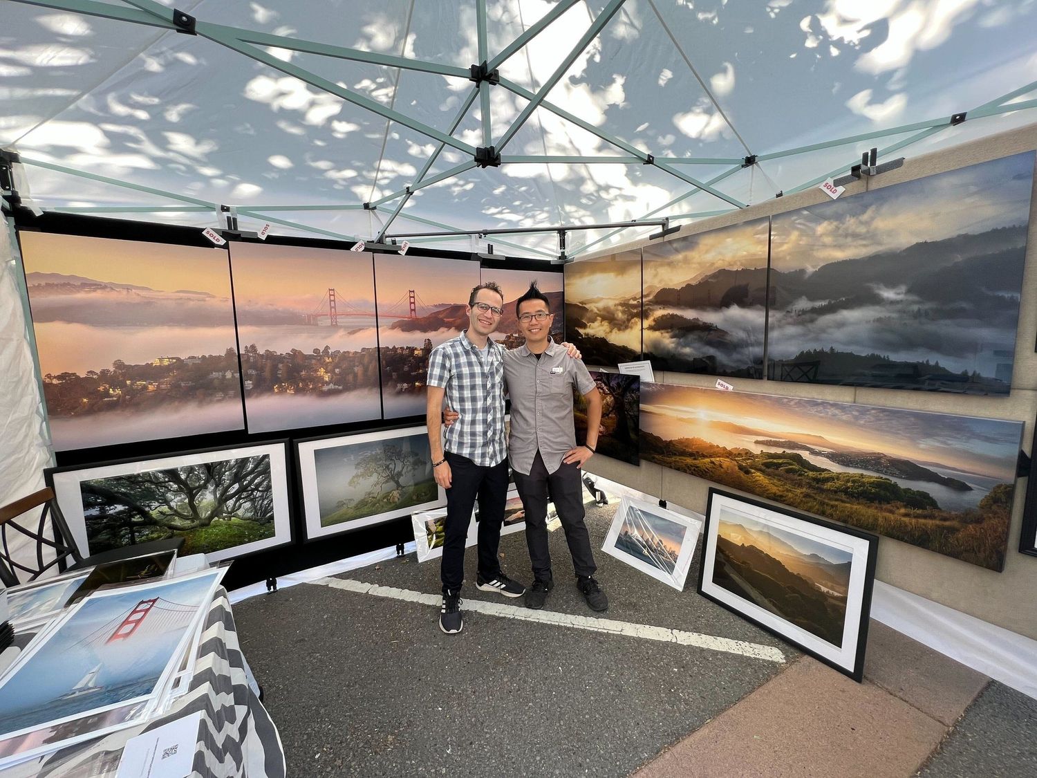 An art booth at a fair displays large landscape photography prints against a white tent backdrop during sunset.
