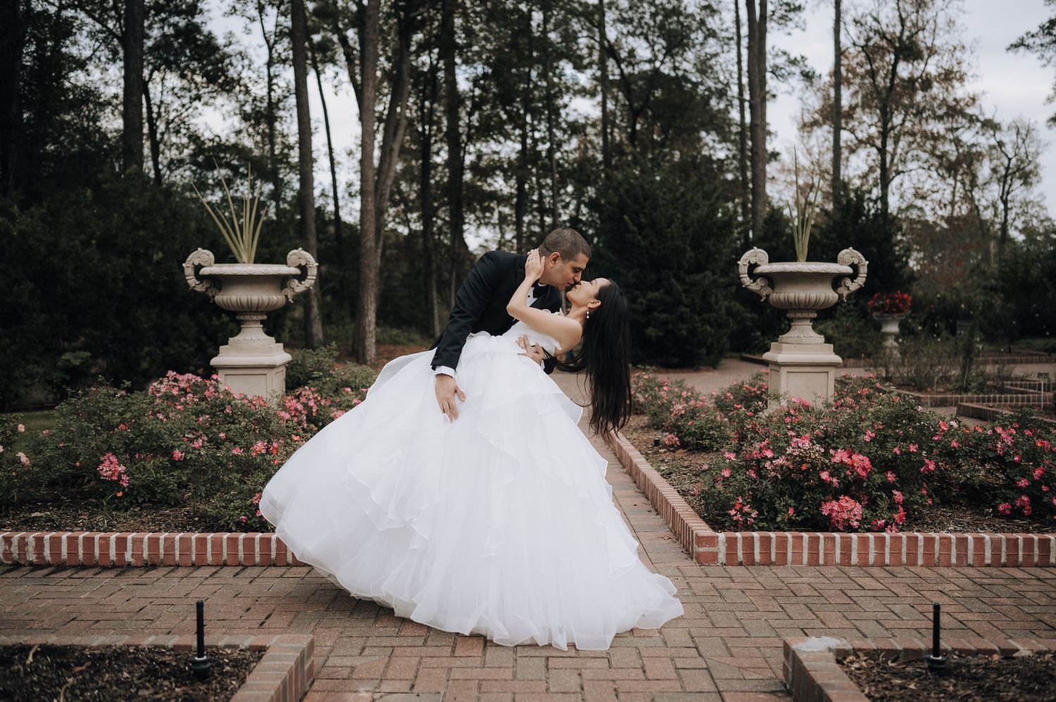 A dramatic wedding pose on brick pavers between ornate garden urns with roses in the background.