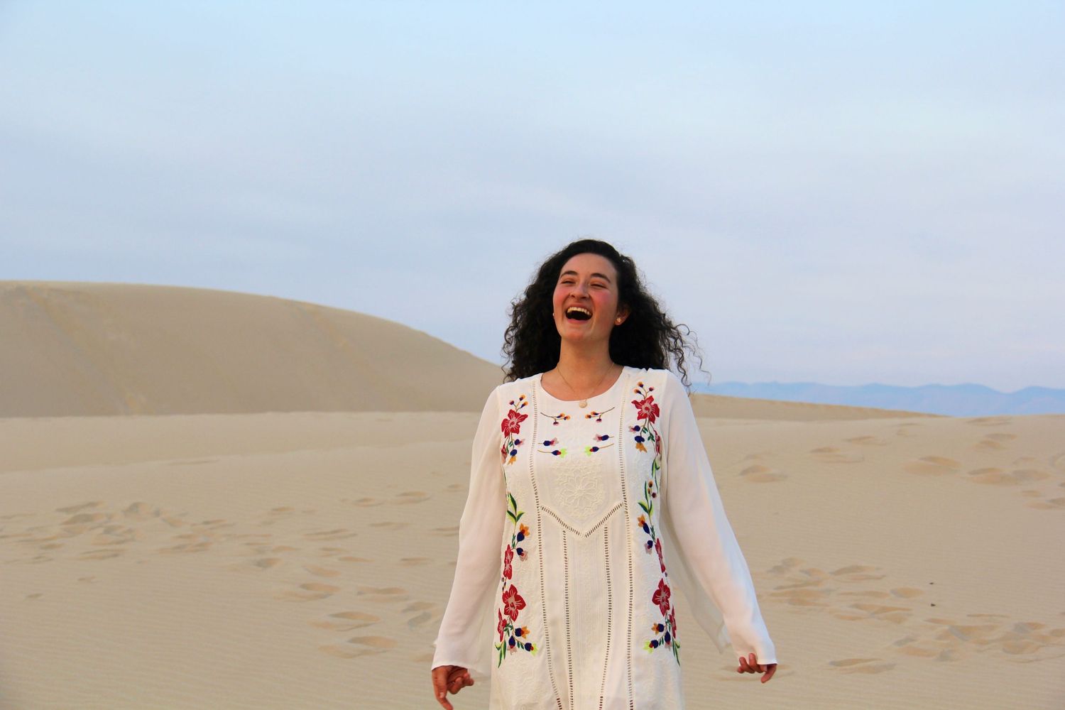 Woman in white embroidered dress laughing joyfully while standing on sand dunes in desert landscape.