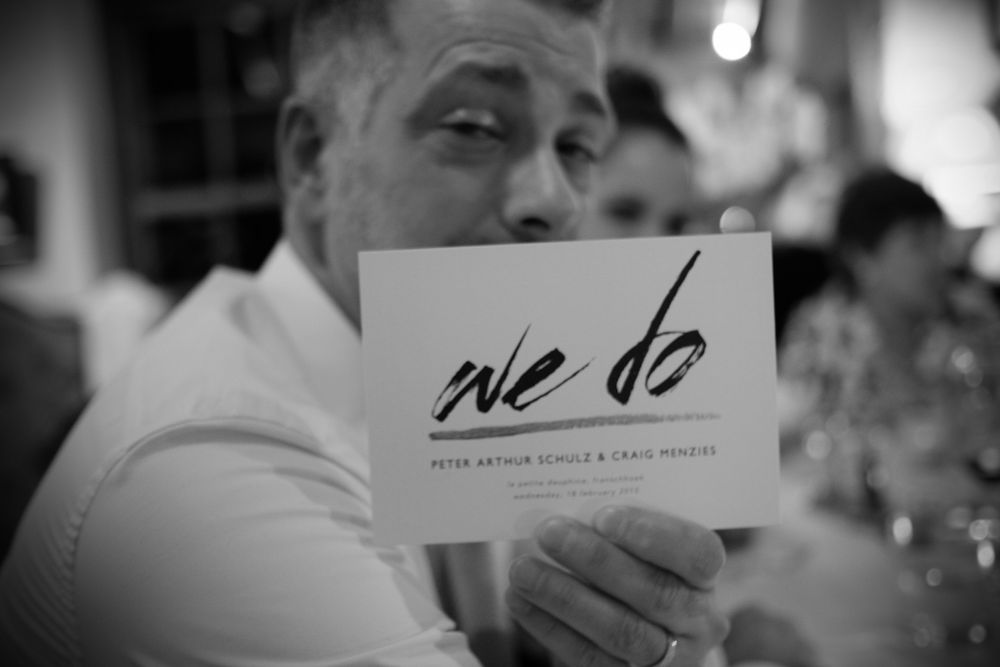 Older man holding a black and white wedding card that reads we do at a reception dinner.