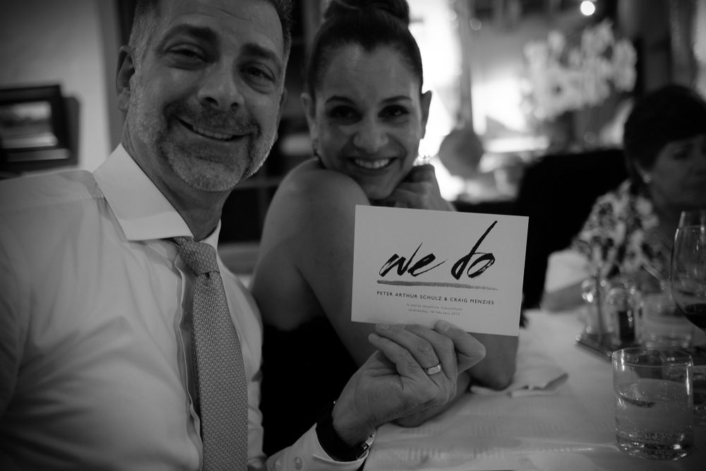 Smiling couple holding a we do wedding card together at an elegant dinner table.