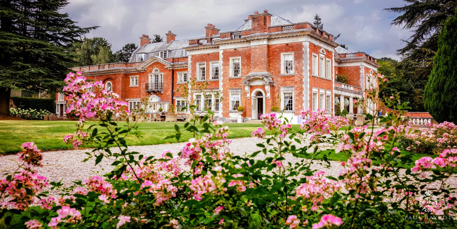 Grand red brick mansion with manicured gardens and blooming pink roses in the foreground.