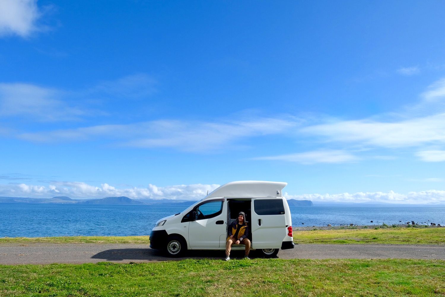 A white van parked along a scenic coastal road with blue ocean and mountains in view.