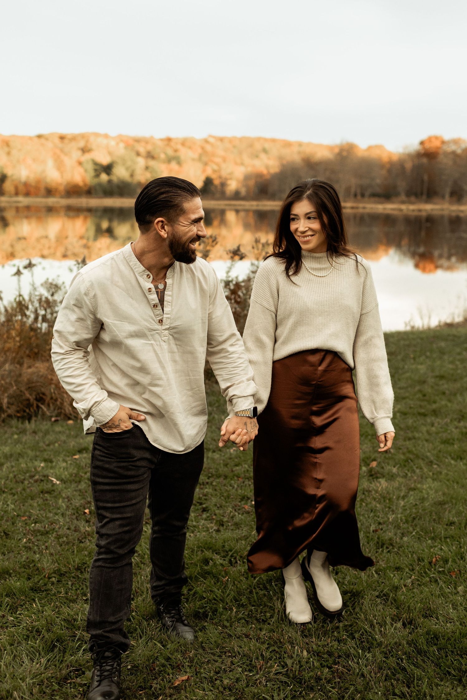 Couple in autumn attire holding hands while walking by lakeside during golden hour.