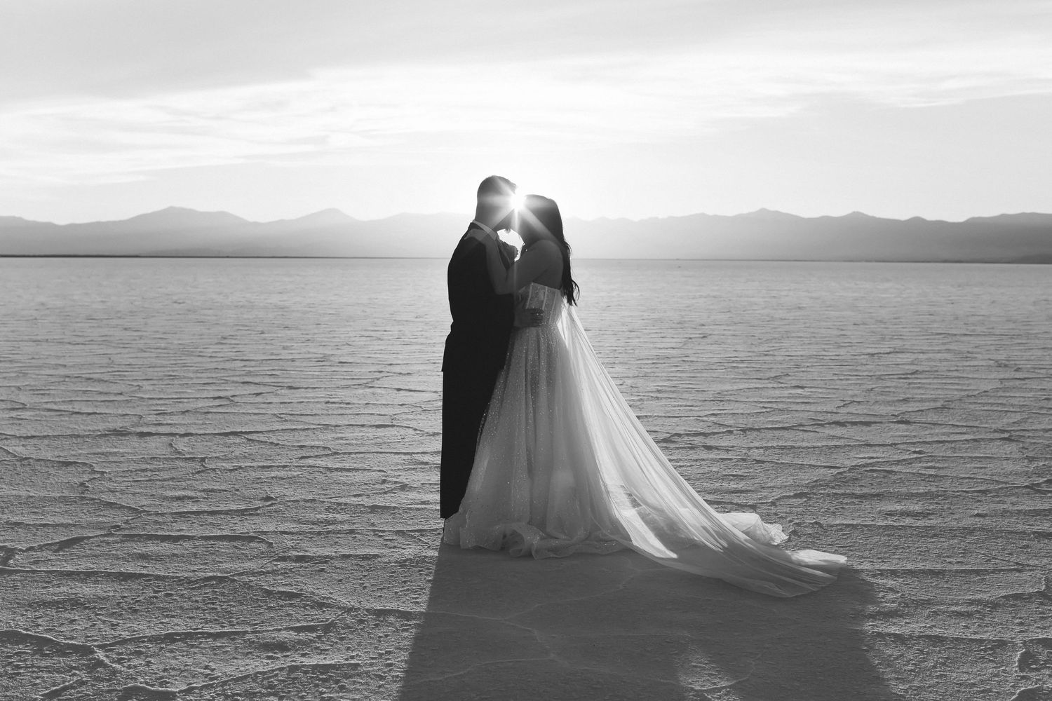 Wedding couple embraces on salt flats with mountains and cloudy sky in monochrome.