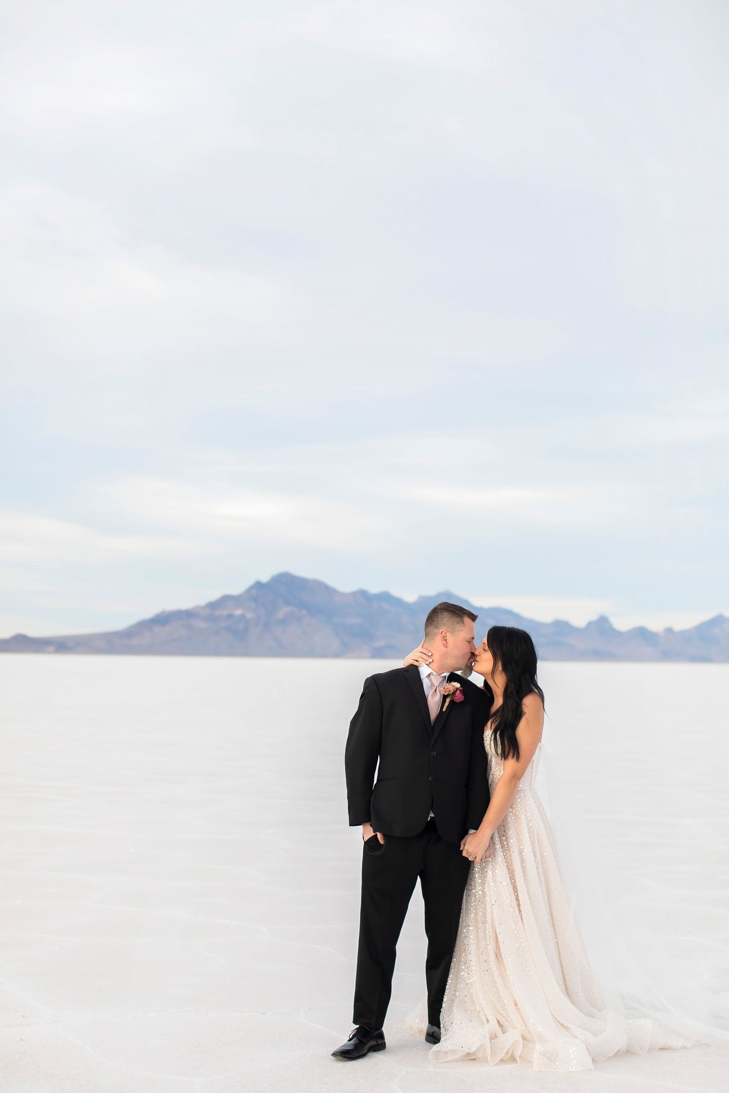 Wedding couple standing together on pristine white salt flats with mountain backdrop.