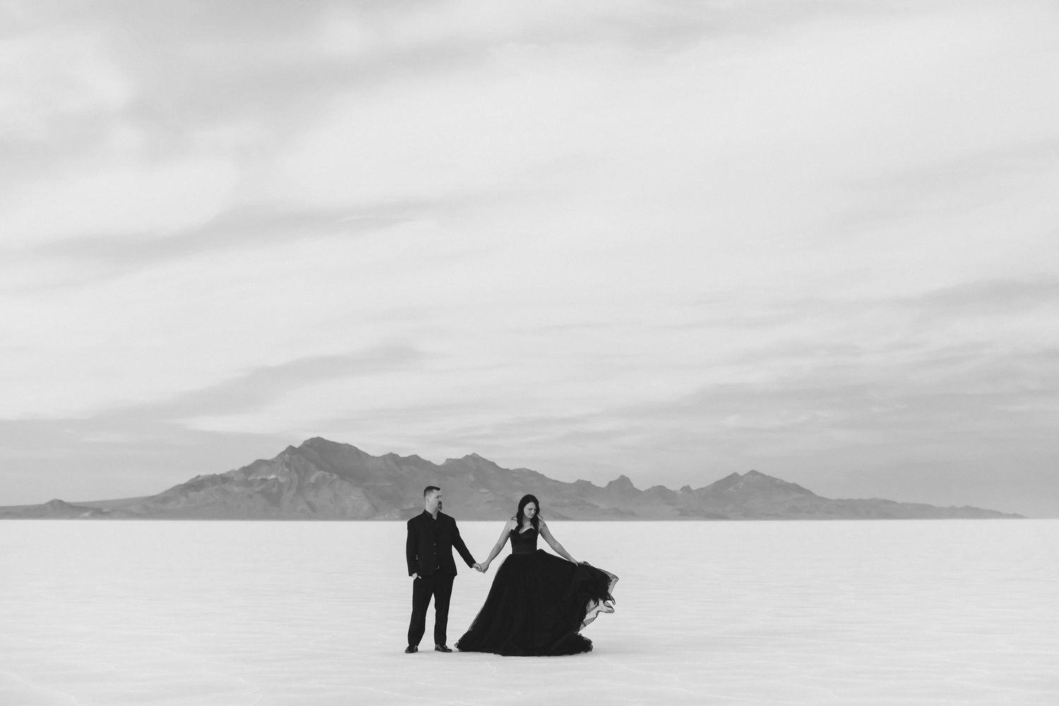 Couple in formal attire stand dramatically on salt flats with mountain backdrop in black and white.