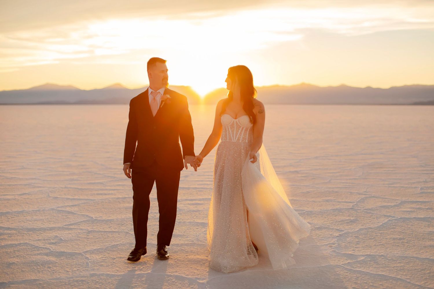 Couple in wedding attire walking on salt flats during golden hour.