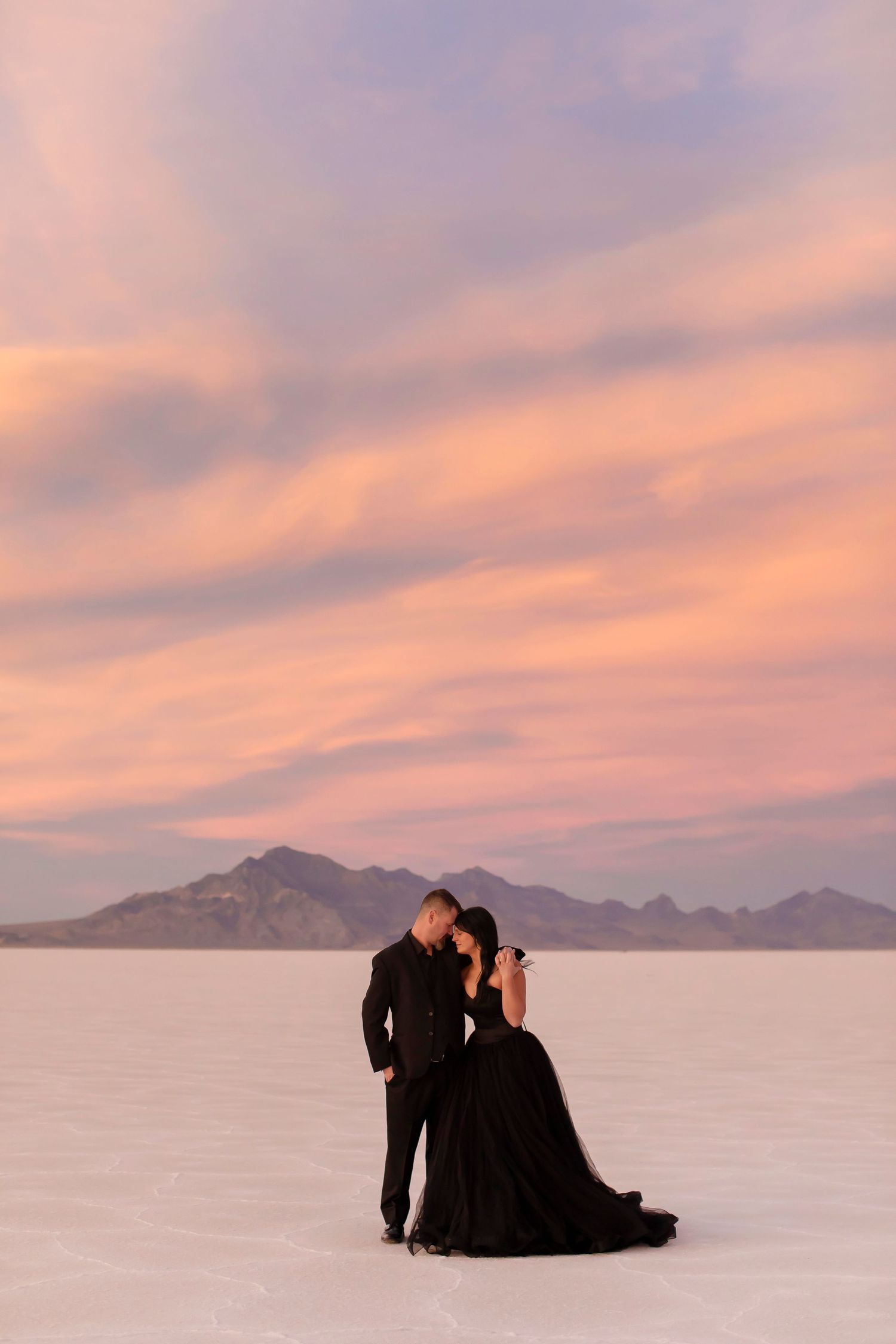 Couple in formal wear pose against pink sunset sky on salt flats with mountain backdrop.