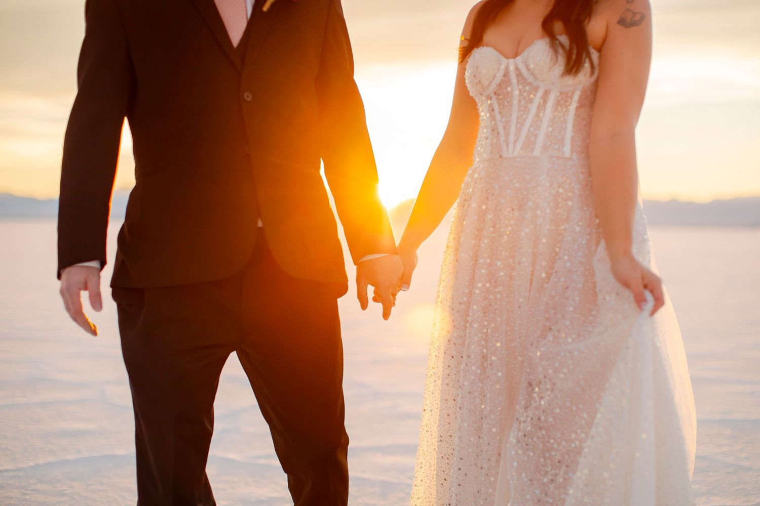 Bride in white dress and groom in suit holding hands at sunset by water.