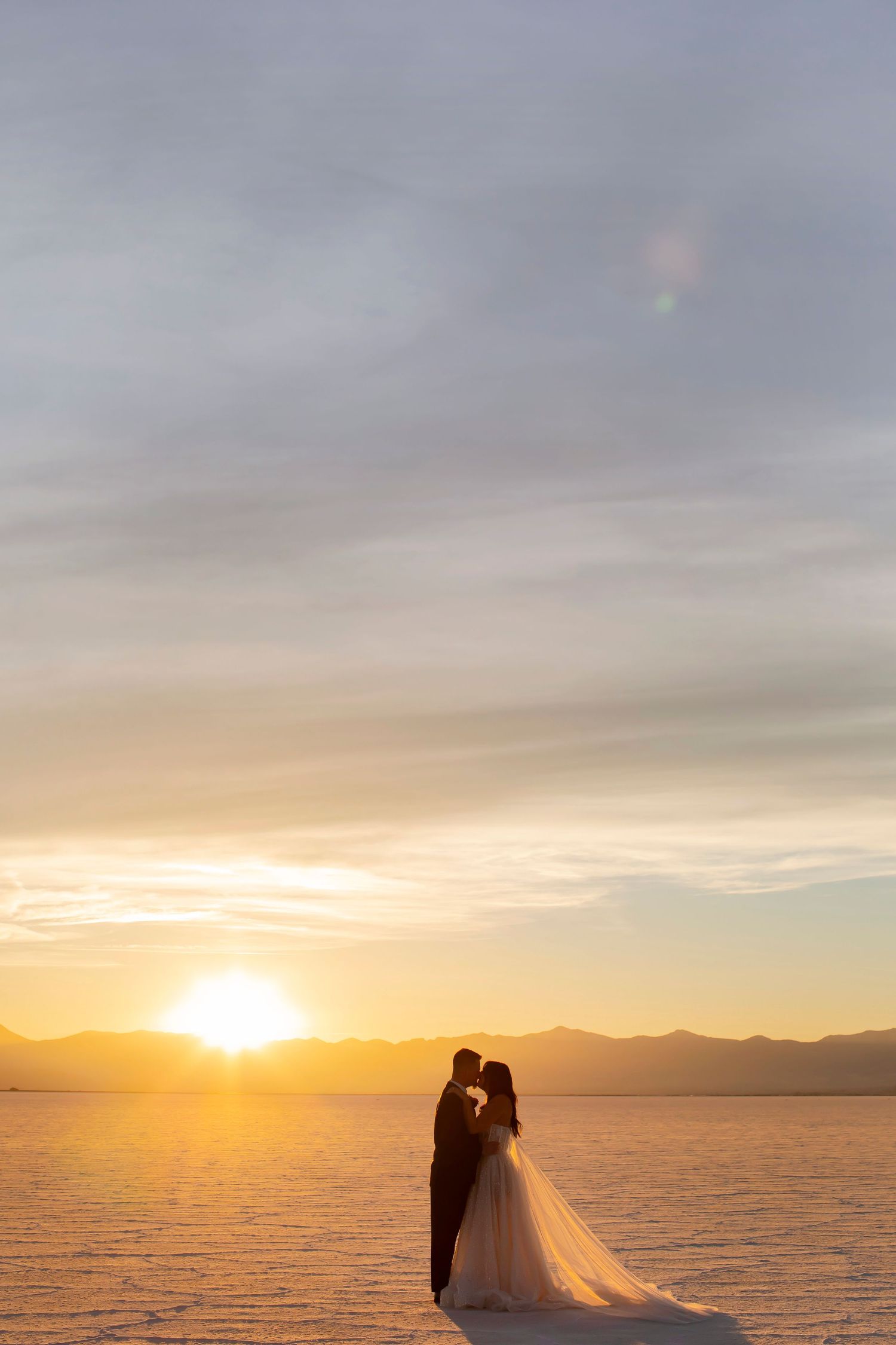 Silhouetted couple in formal attire sharing an intimate moment at sunset on salt flats with mountains in background.