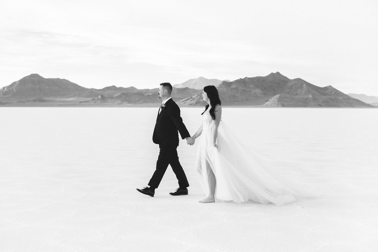 Bride and groom walk hand in hand across white salt flats in black and white photography.
