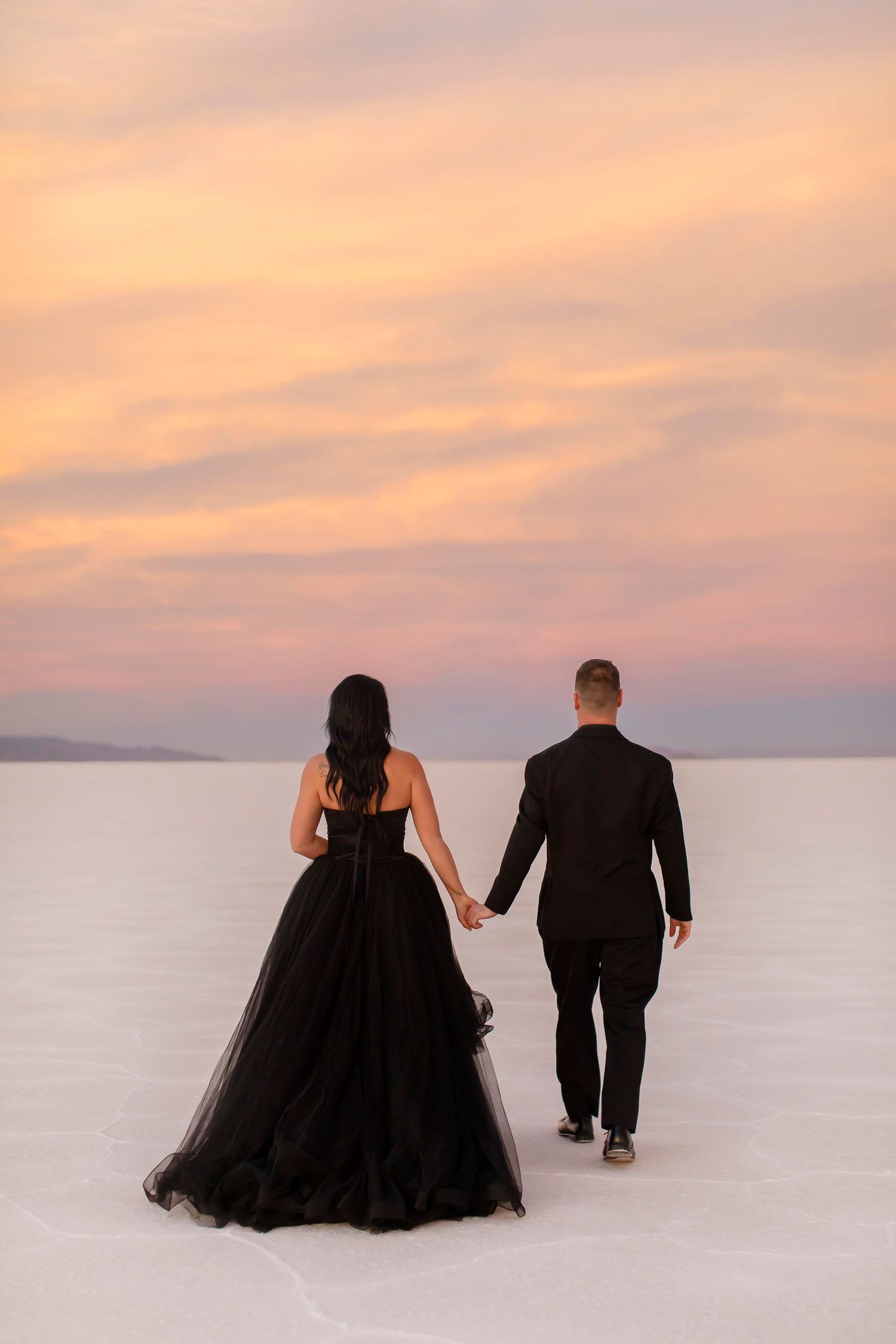 Couple walking hand in hand on salt flats during sunset wearing formal black attire.