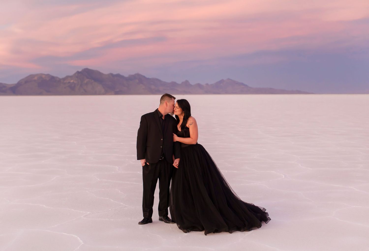 Romantic couple in formal wear sharing intimate moment on salt flats at sunset.
