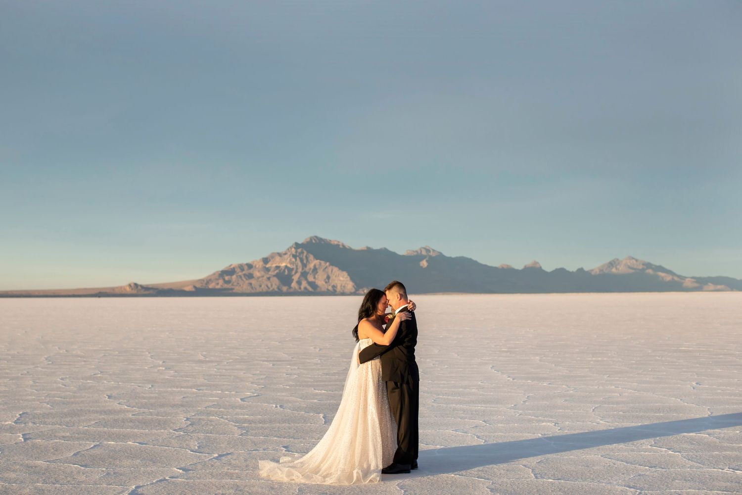 Wedding couple embracing on white salt flats with mountain range backdrop under blue sky.