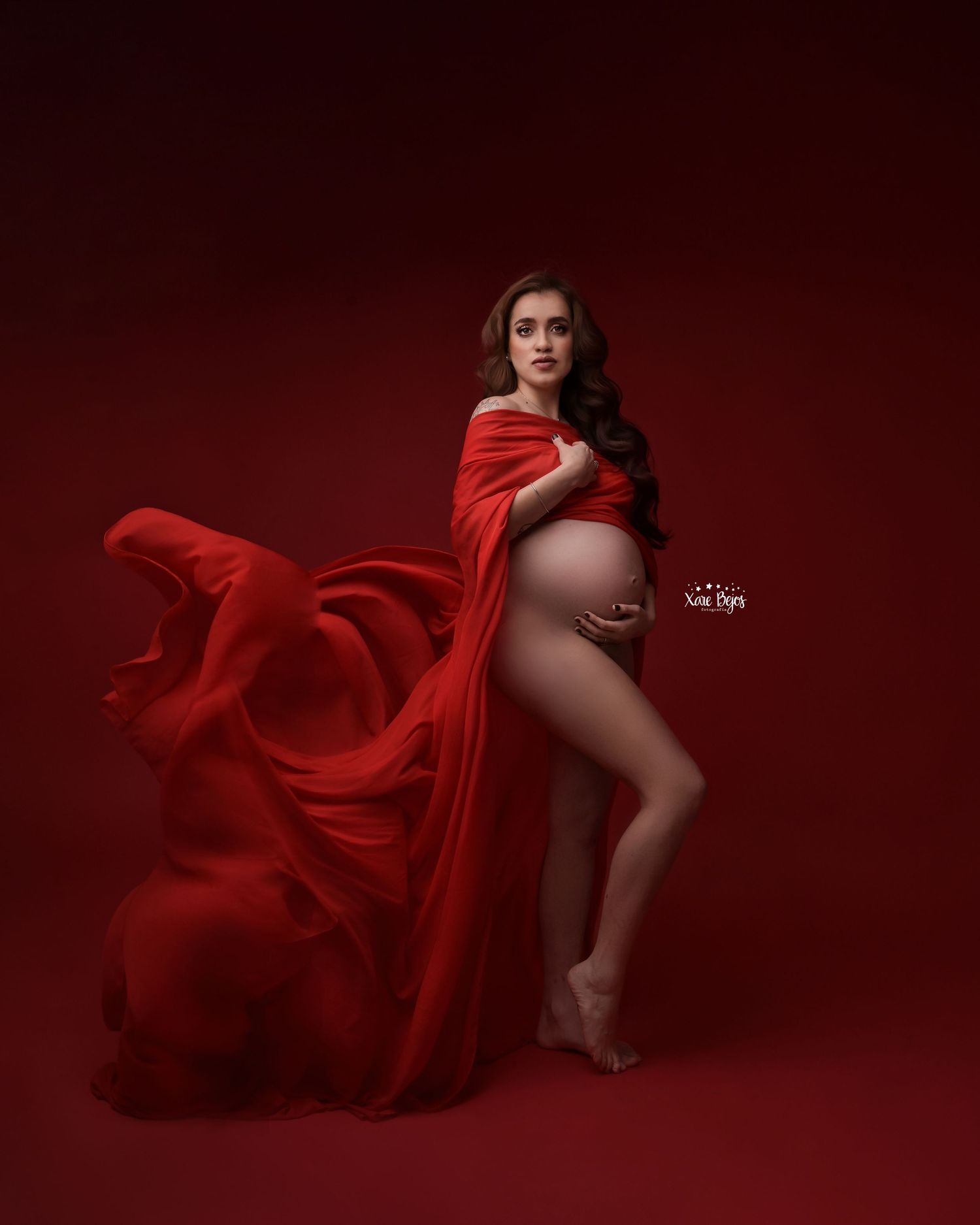 Dynamic maternity portrait with flowing red fabric against deep red background.