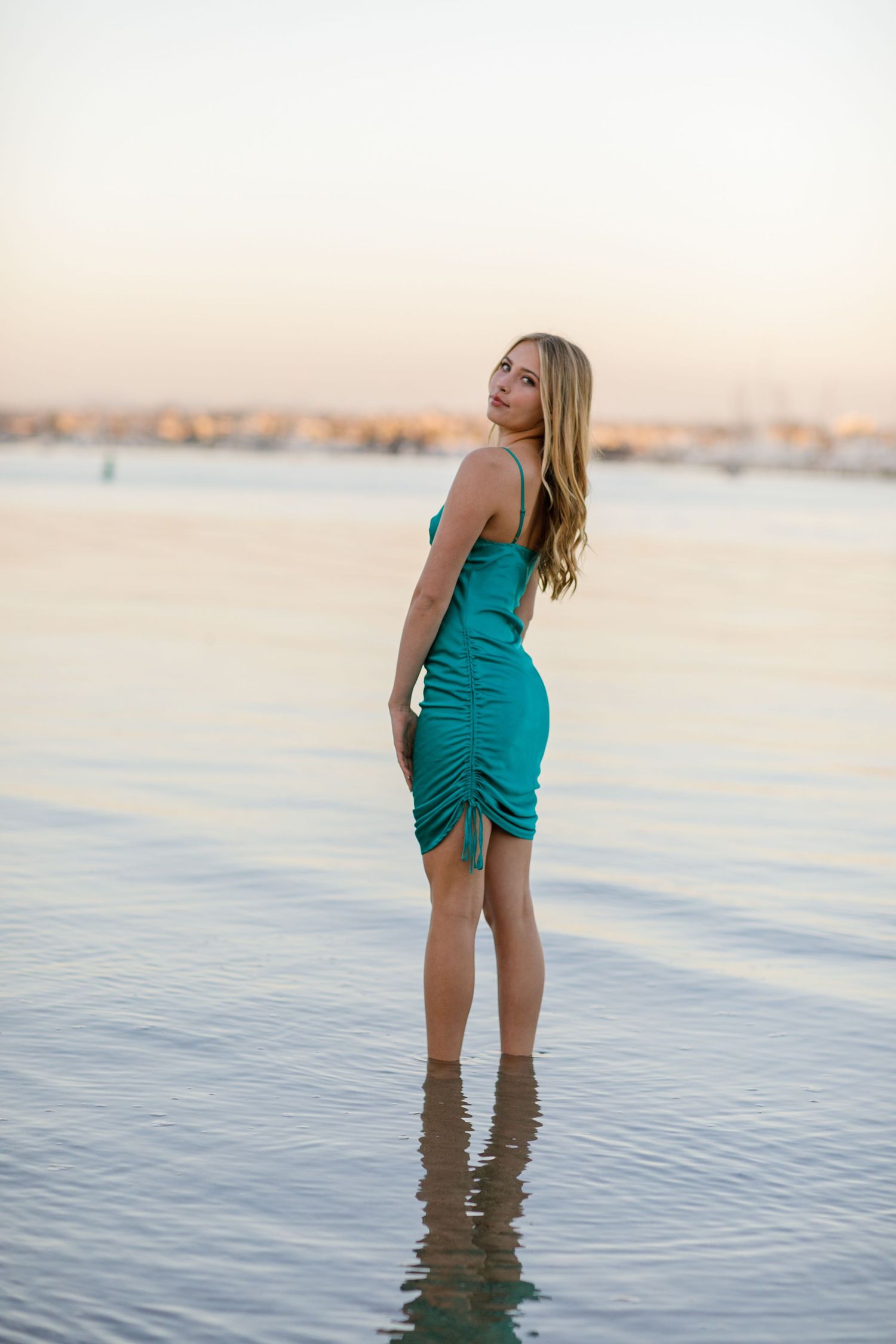 A high school senior in a turquoise dress stands in shallow ocean water at sunset as gentle waves lap at their feet.