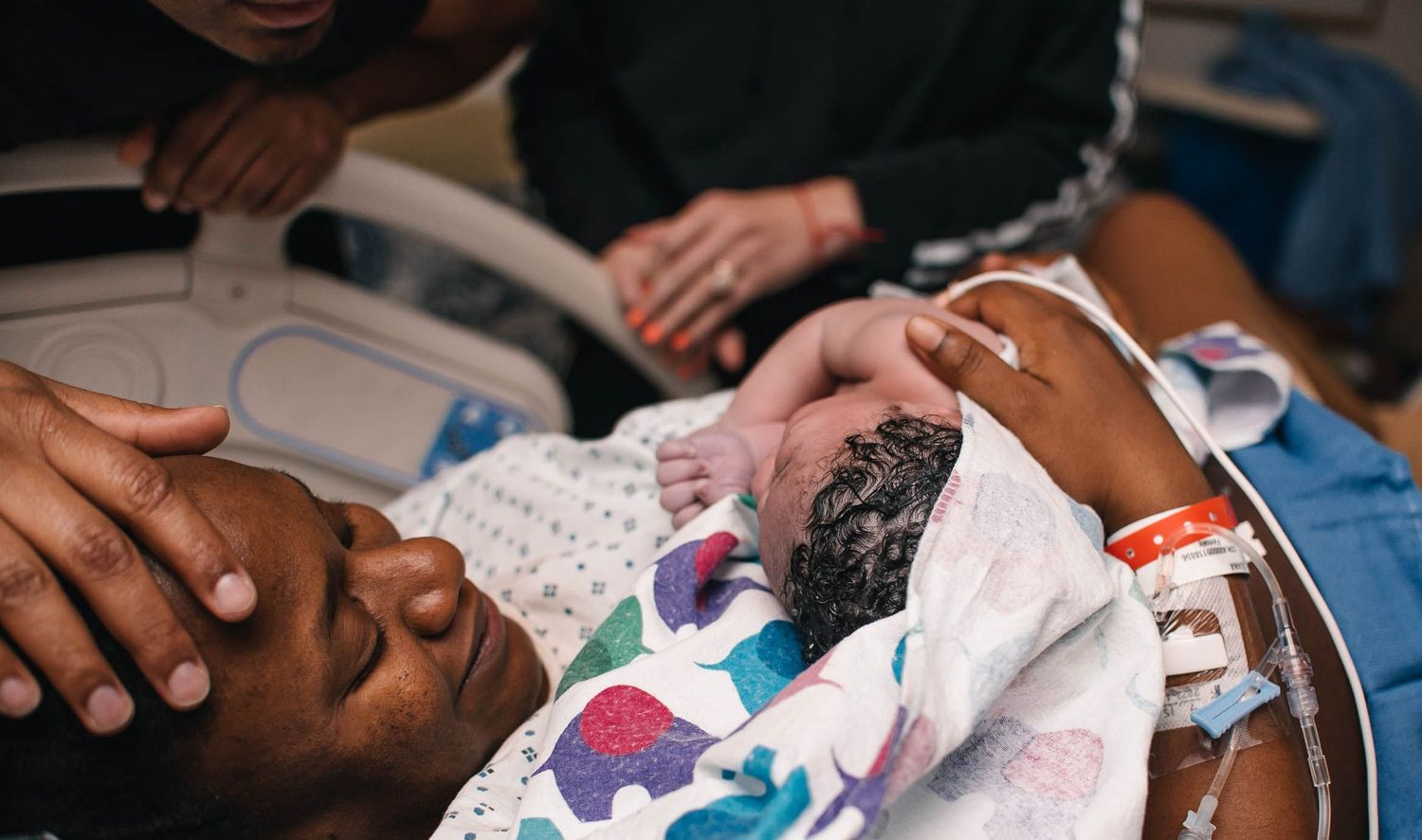 lady smiling while holding a newborn baby with a man's hand on her forehead lying in a blanket in a hospital bed
