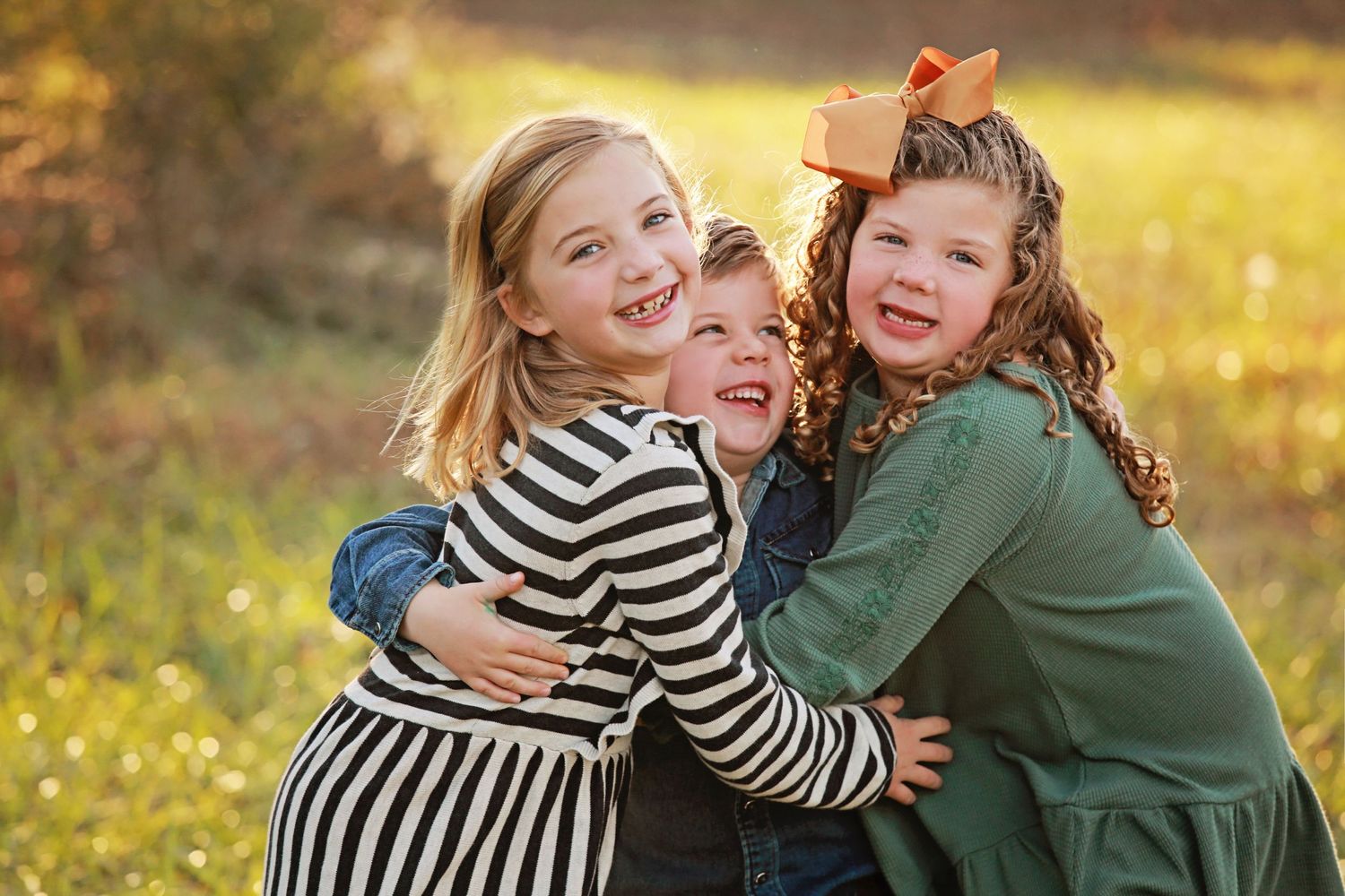 Three children embracing and laughing together in golden sunlit field with yellow wildflowers.