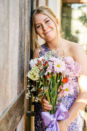 Bridesmaids in purple sequin dresses hold colorful flower bouquets while posing against a rustic wooden wall.