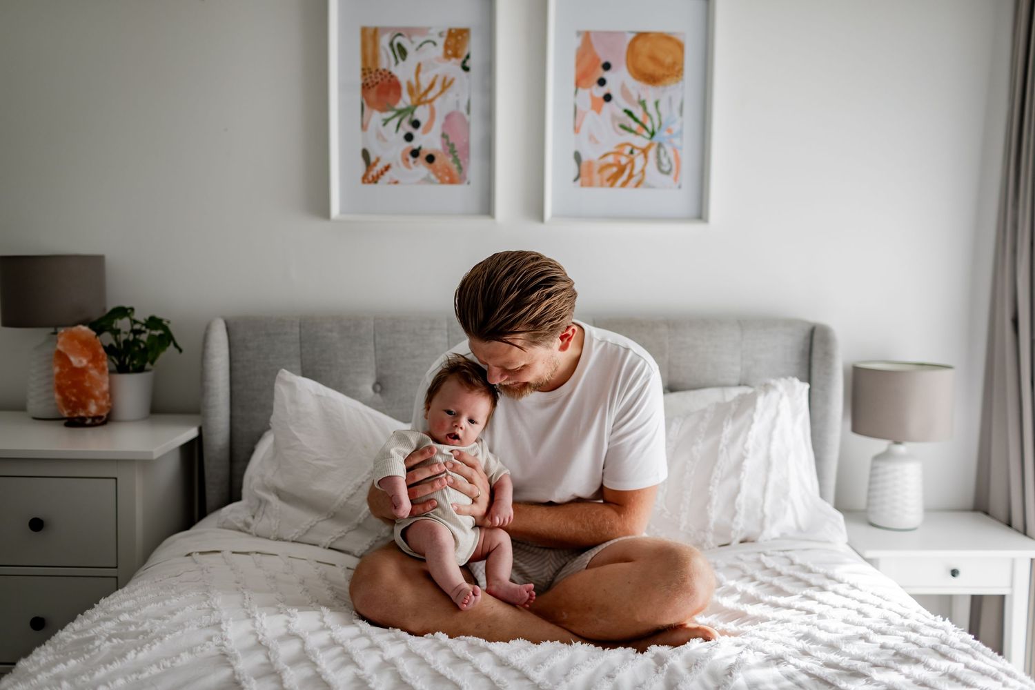 Tender moment between parent and baby on white bedding with abstract art on wall.