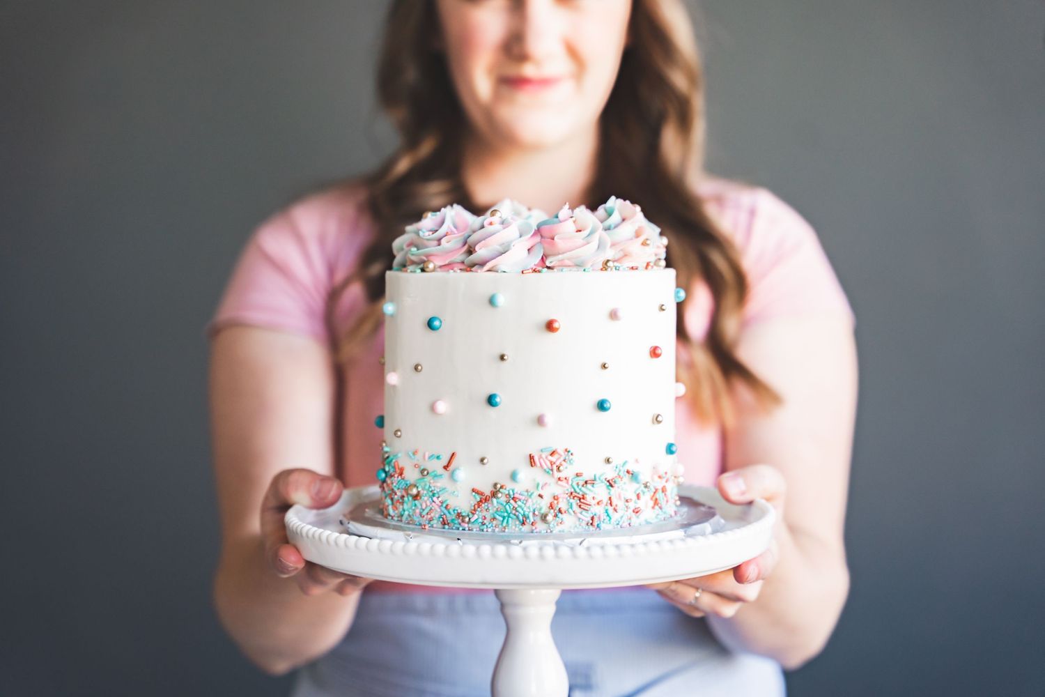A decorated white cake with pastel polka dots and sprinkles being held on a cake plate.