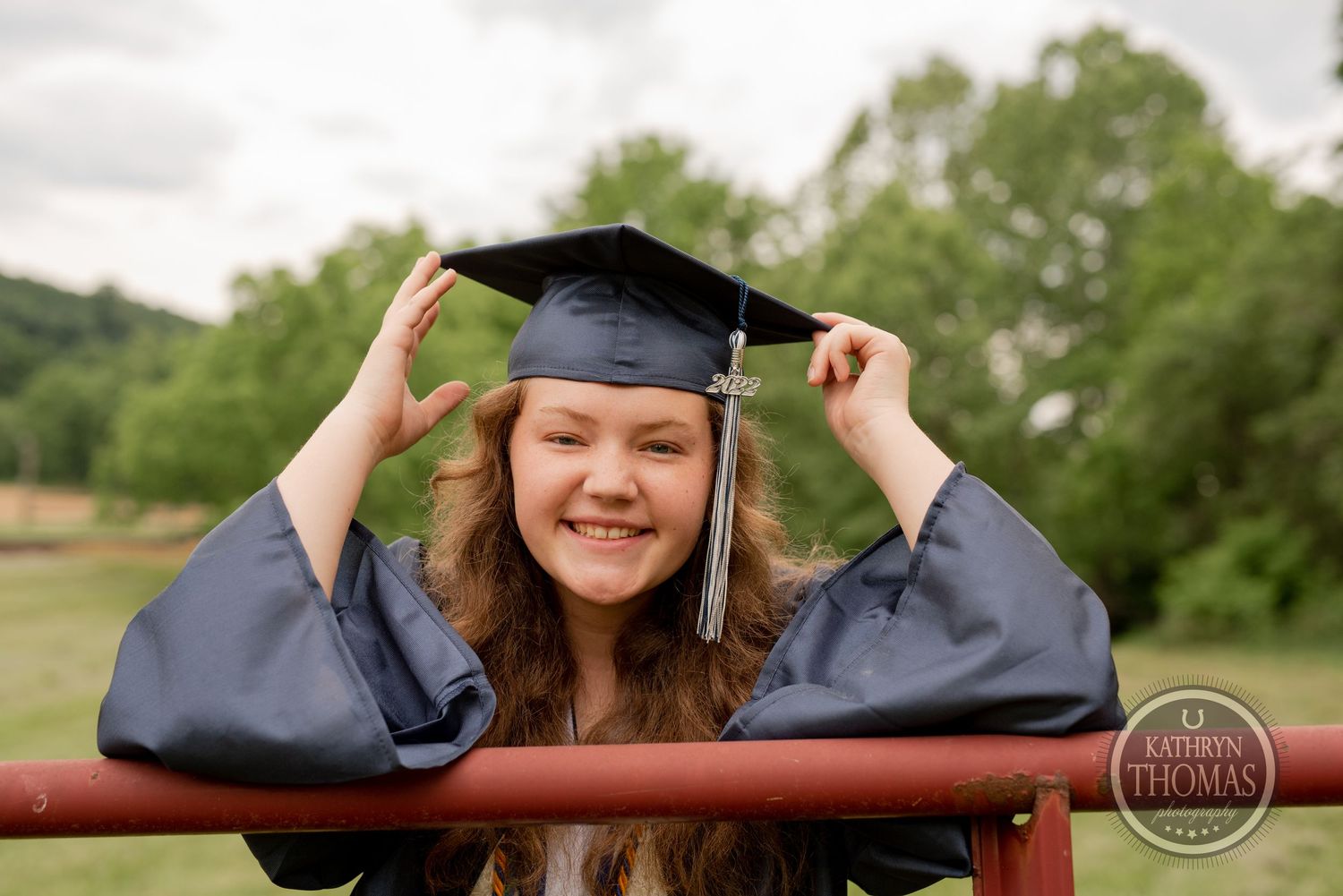 Graduate adjusts mortarboard cap while smiling and leaning on red railing outdoors.