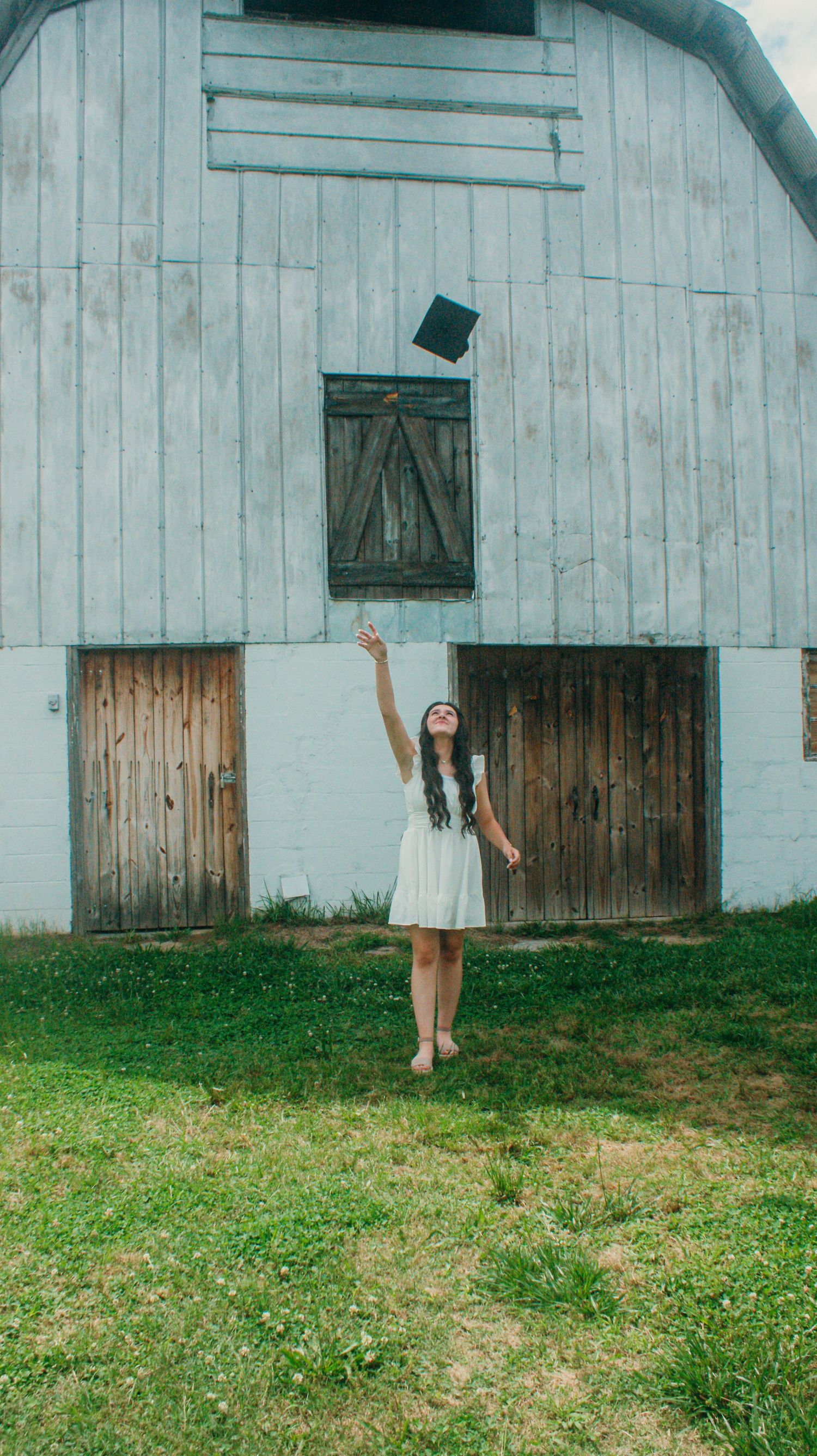 A graduate in white dress tosses cap in front of rustic white barn on grassy field.