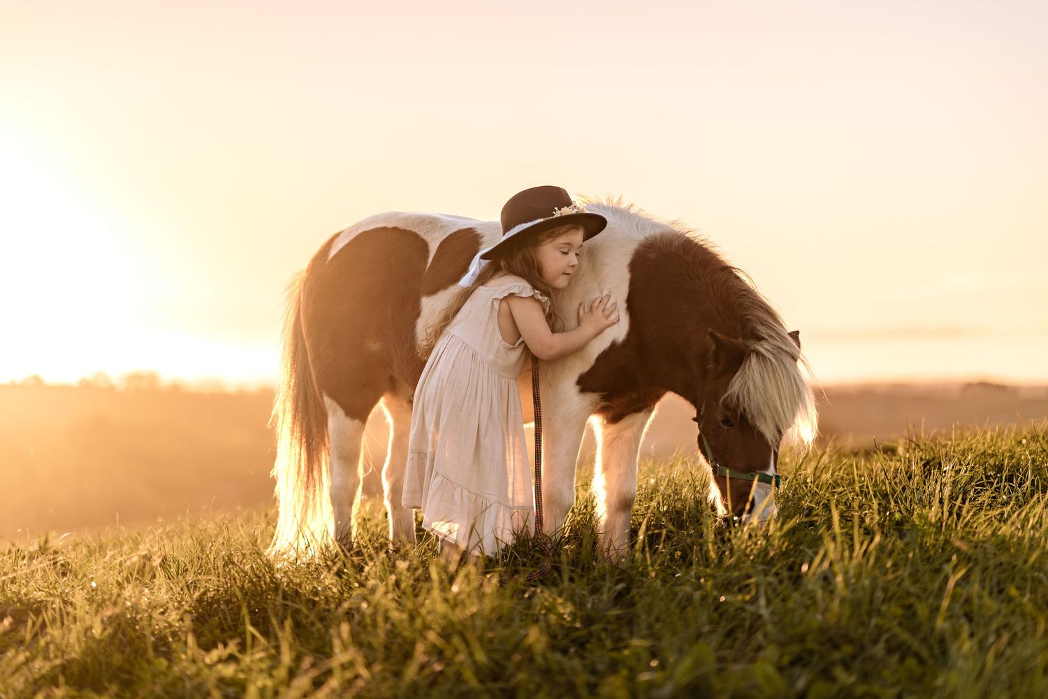 A dreamy sunset scene with two horses grazing beside someone in a flowing white dress.