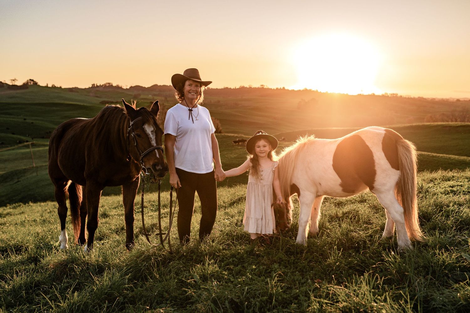 A sunset scene with horses and people standing in a grassy field.