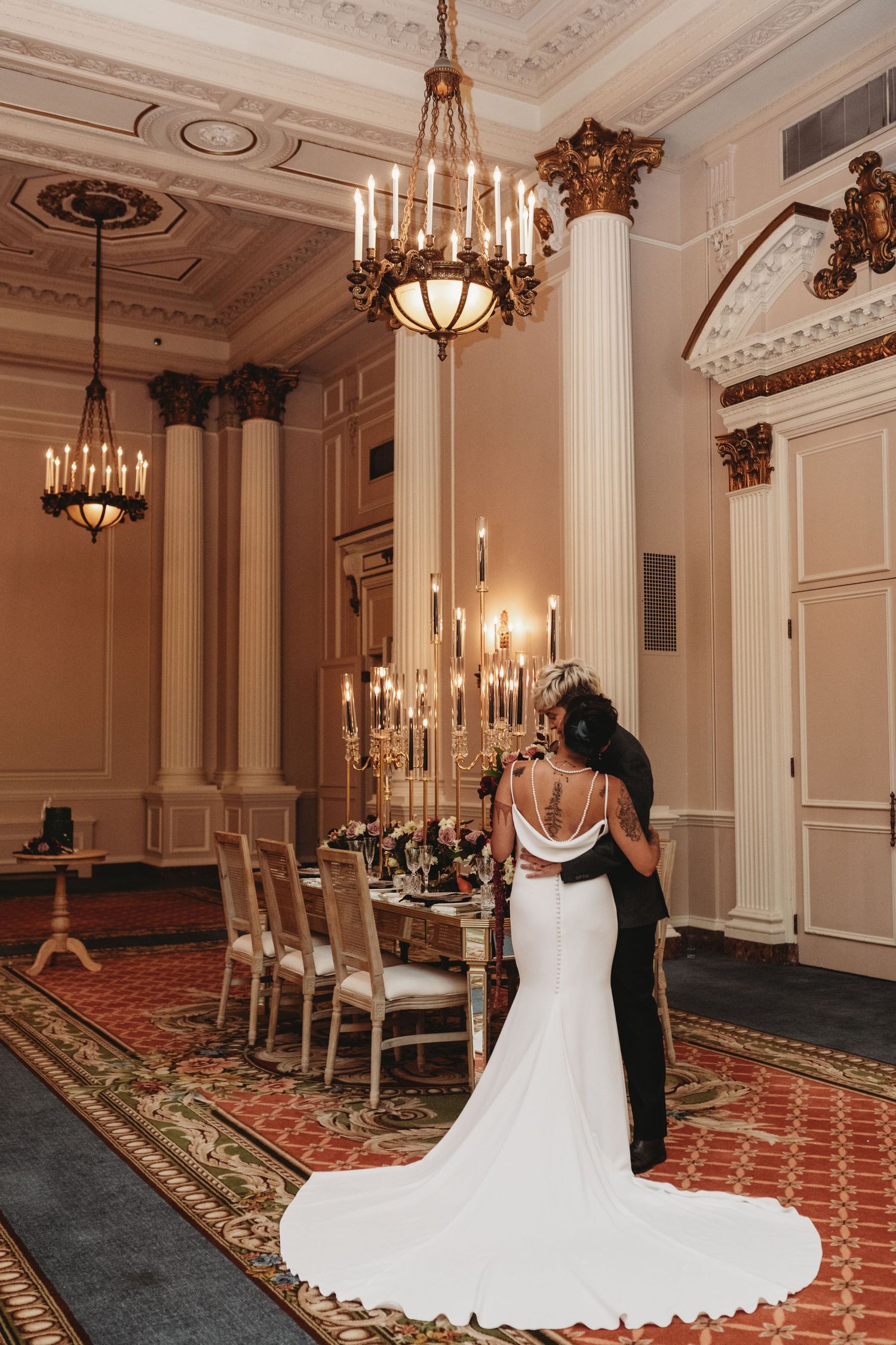 A couple in wedding attire stands in an elegant room with chandeliers and classical columns.