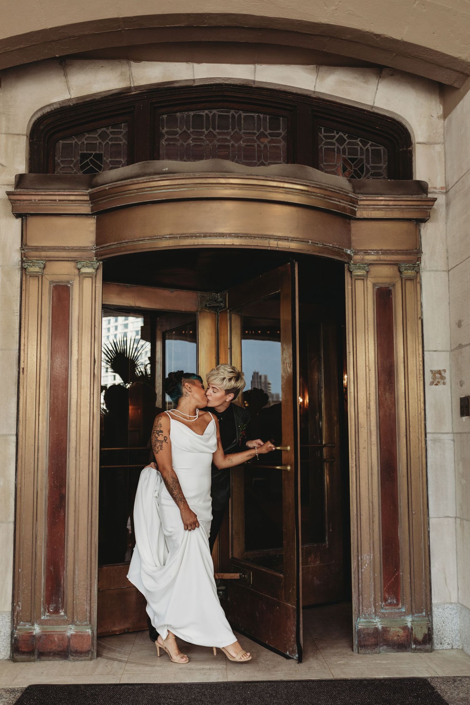 A couple in wedding attire stands in a grand historic entrance with ornate bronze details.