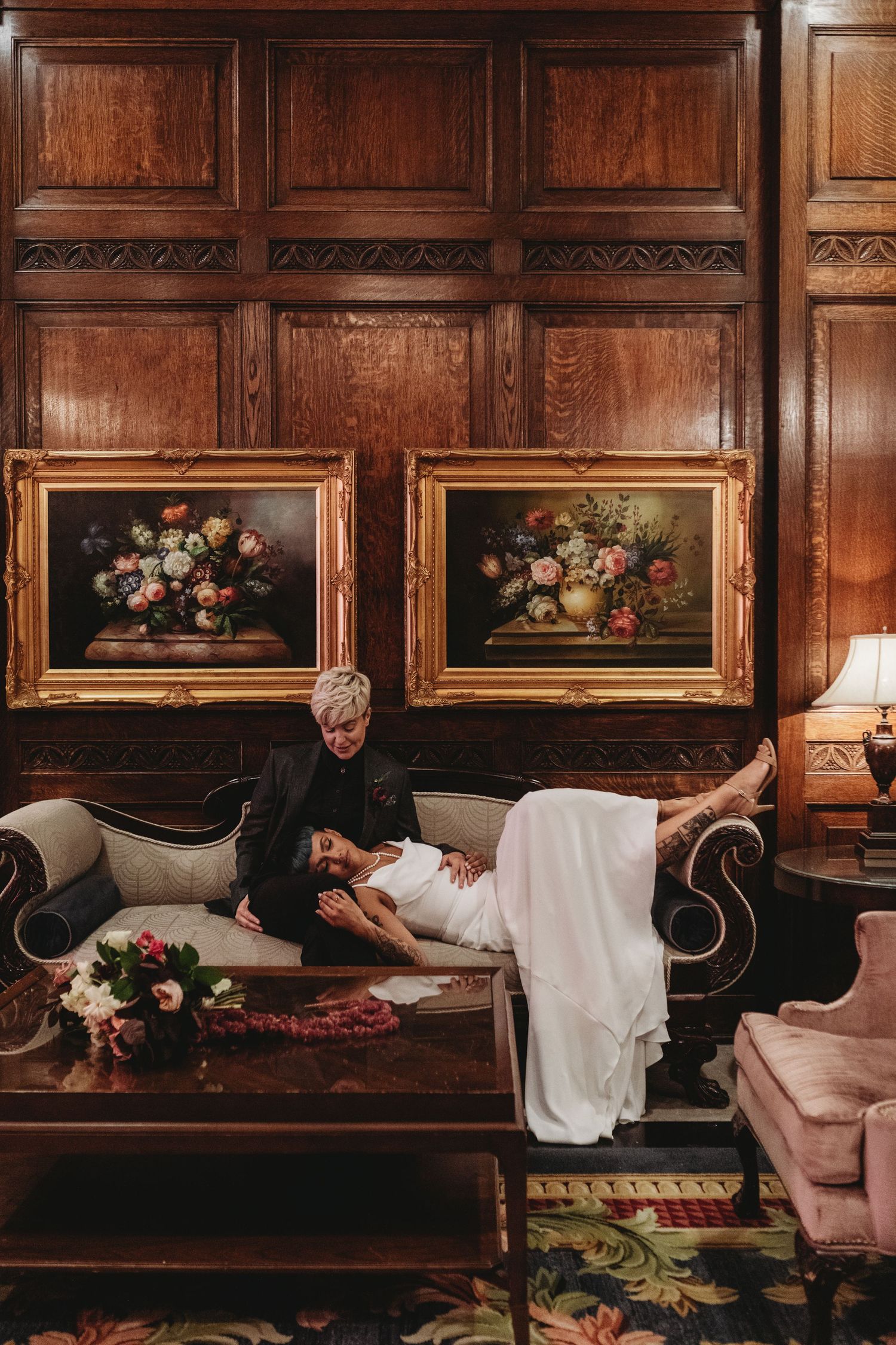 A couple poses on an elegant couch with vintage floral paintings displayed on wood-paneled walls.