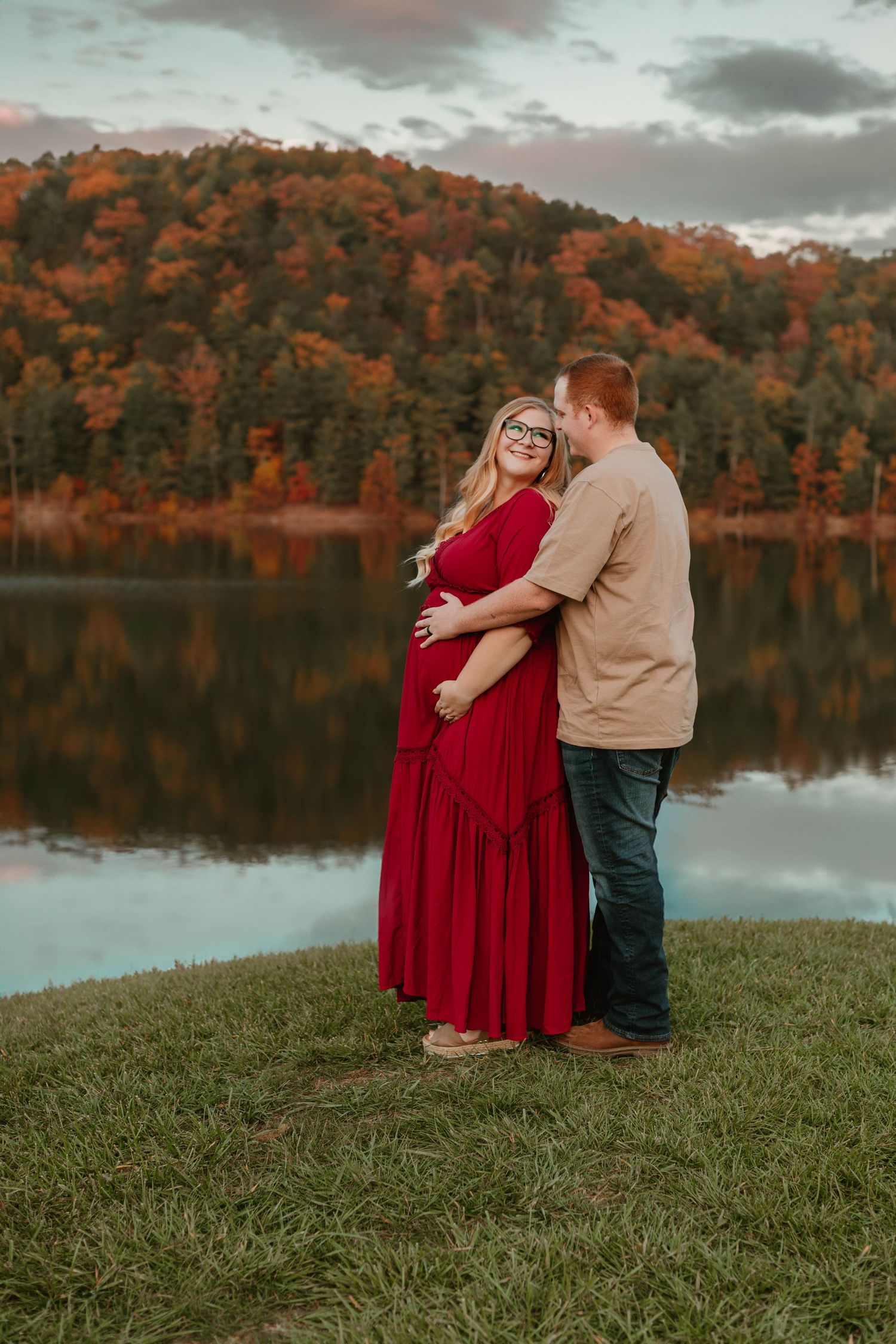 Couple embraces by lakeside during autumn sunset with colorful foliage in background.