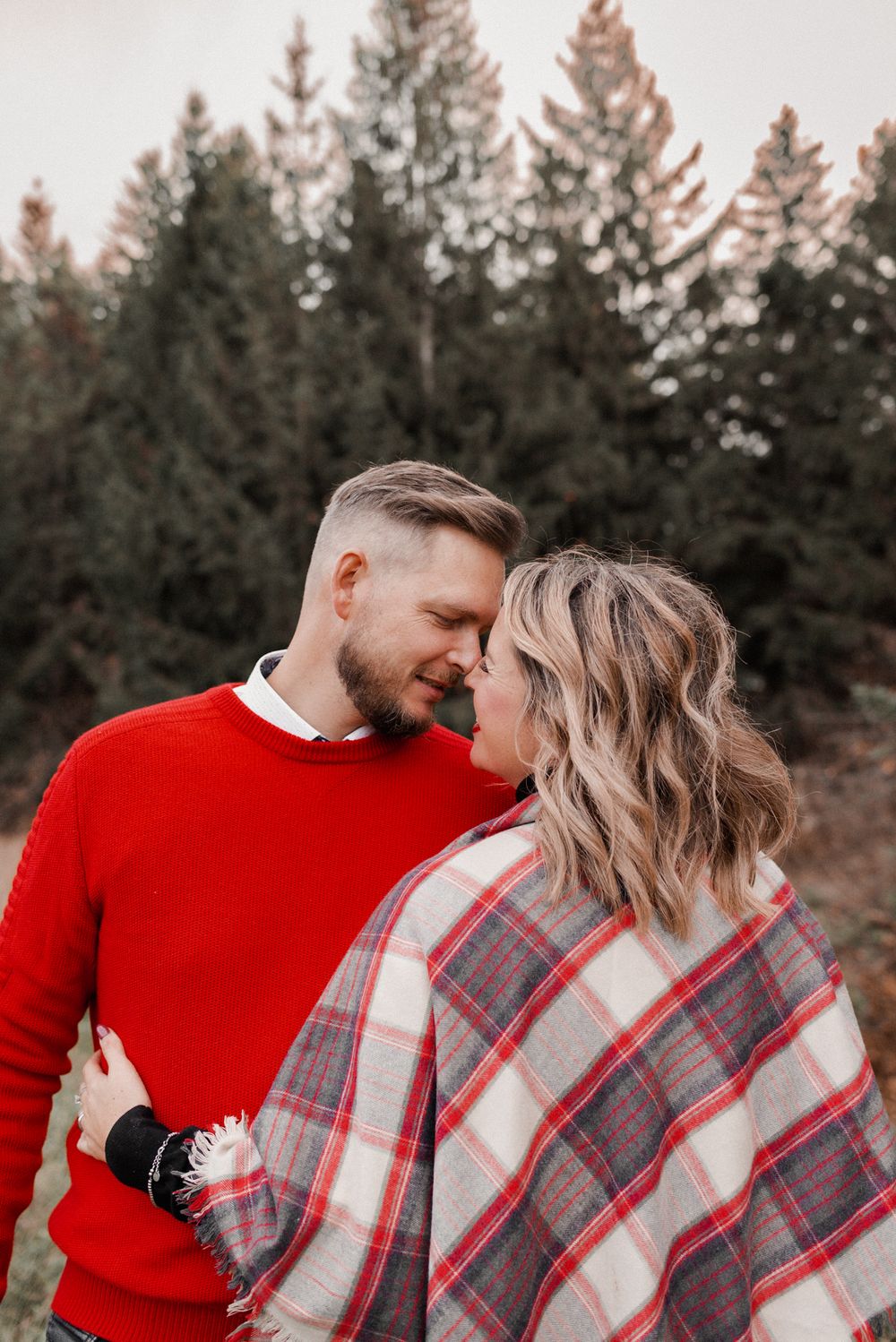 Romantic couple's portrait in forest with red sweater and plaid blanket at sunset.