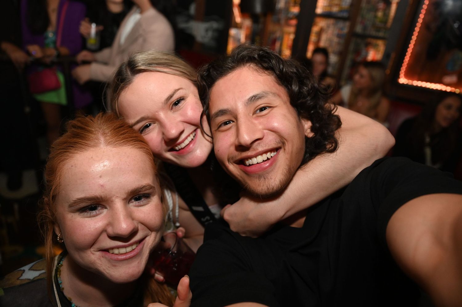 Three friends share laughter and smiles while taking a close-up selfie together at a dimly lit party.