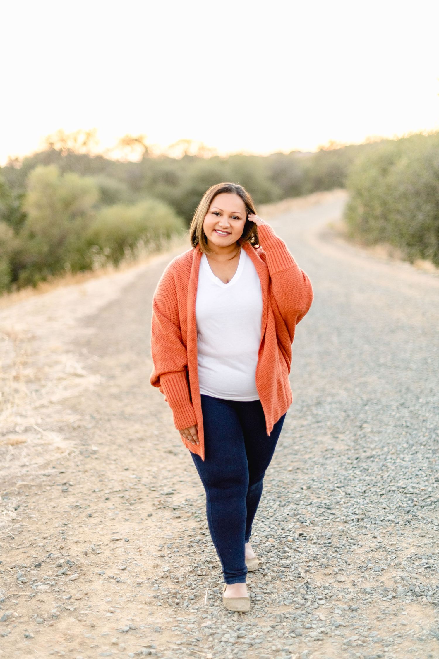 Person in coral cardigan and white top walks along a gravel path during sunset.