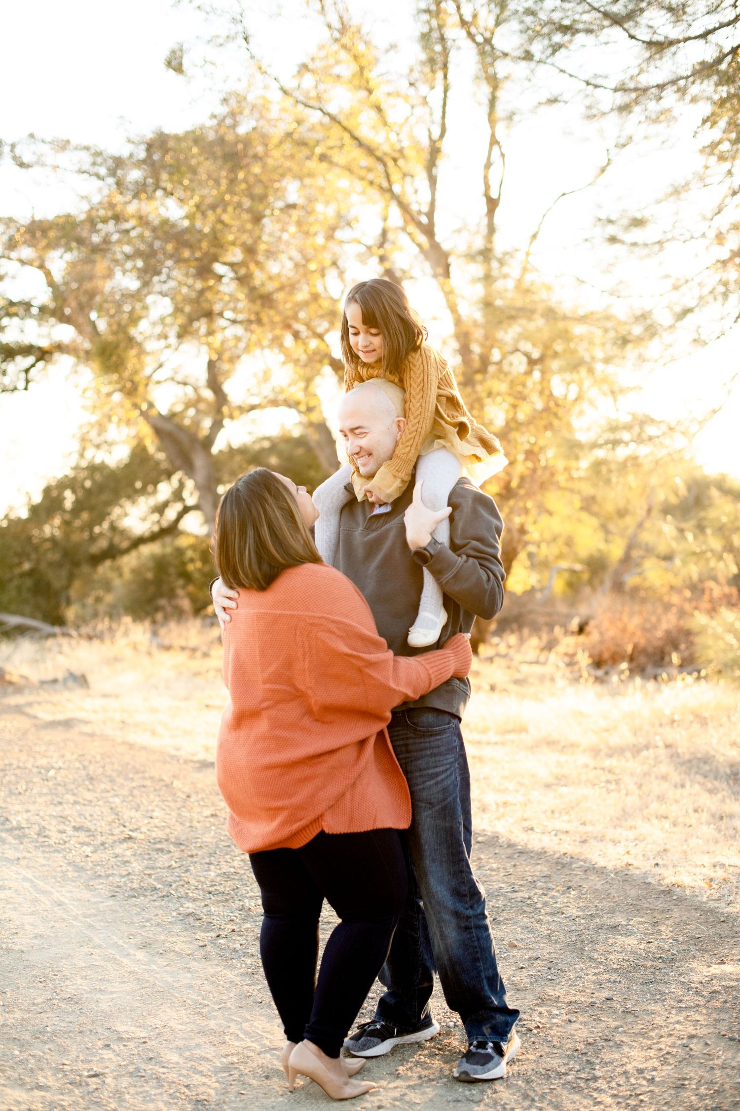 Parents share playful moment with child on shoulders during sunset outdoor photoshoot.