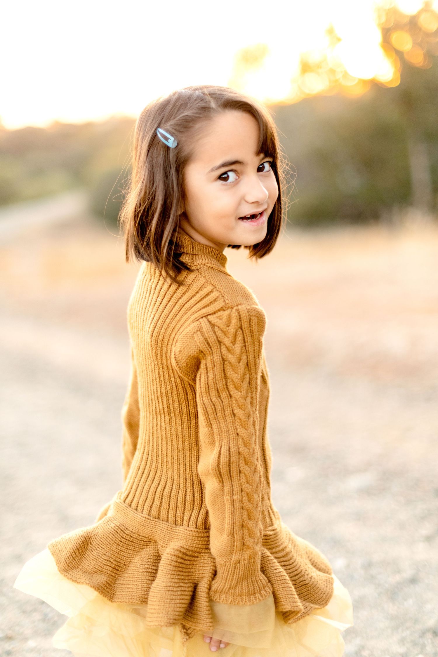Child wearing a cozy yellow sweater poses playfully during an outdoor photo session.