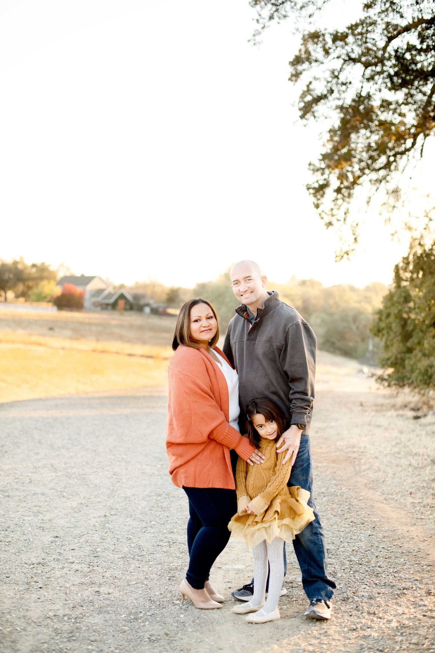Family of three poses together on a dirt path during golden hour sunset.